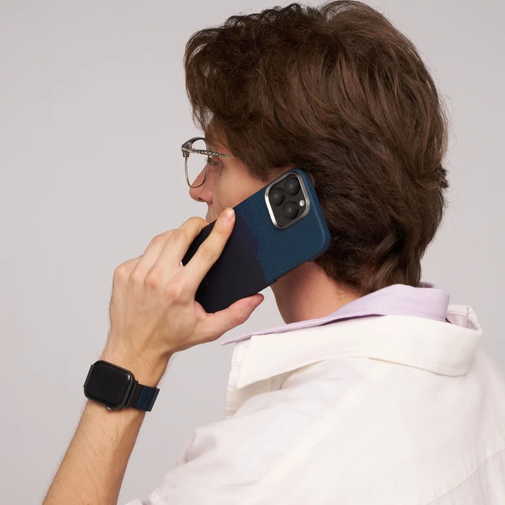 Egy személy telefonál. Szemüveget, fehér inget és okosórát visel. A telefonnak kék tokja van.