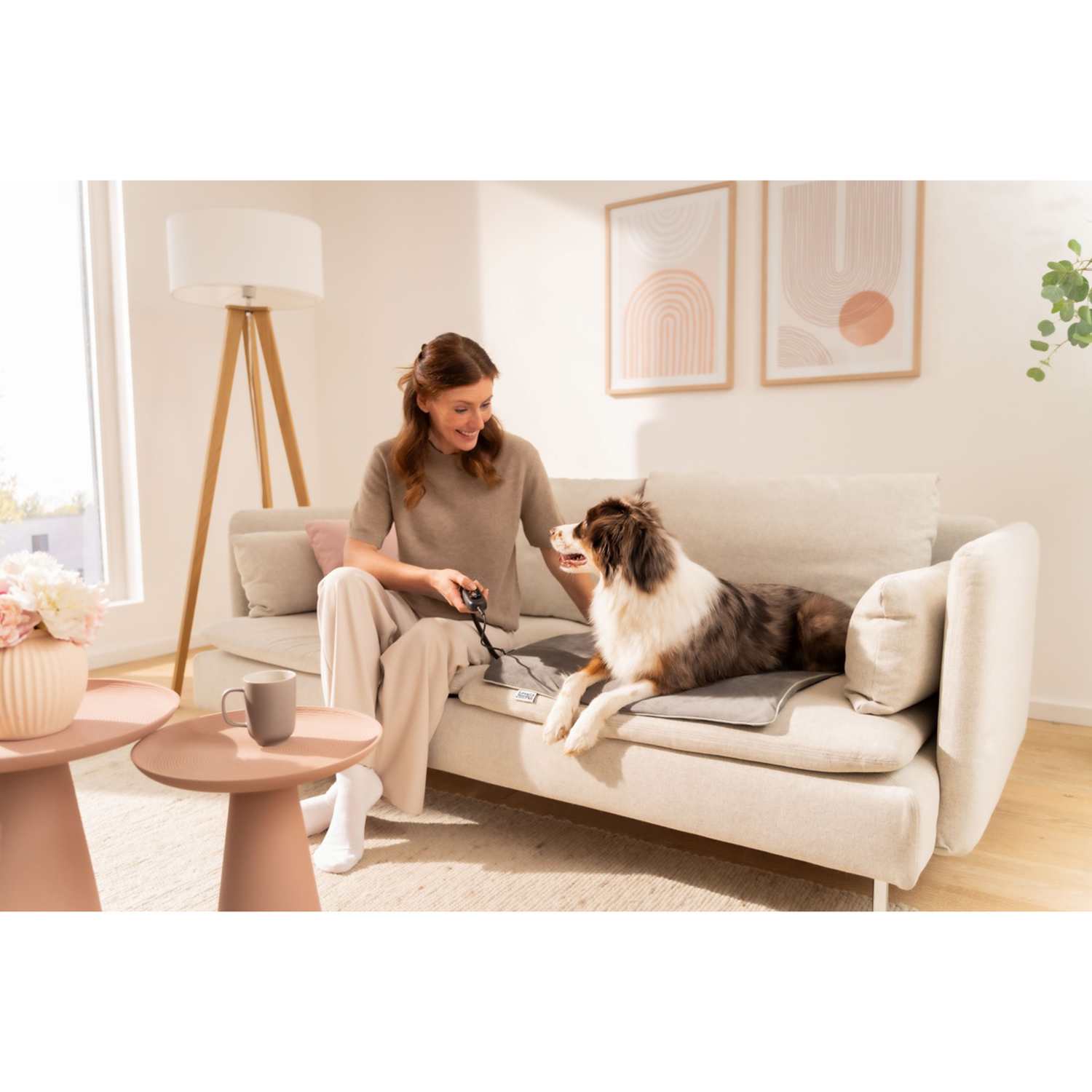 Una donna è seduta su un divano con un cane. Tiene in mano un dispositivo con un cavo. Una lampada e un tavolino con una tazza sono vicini.