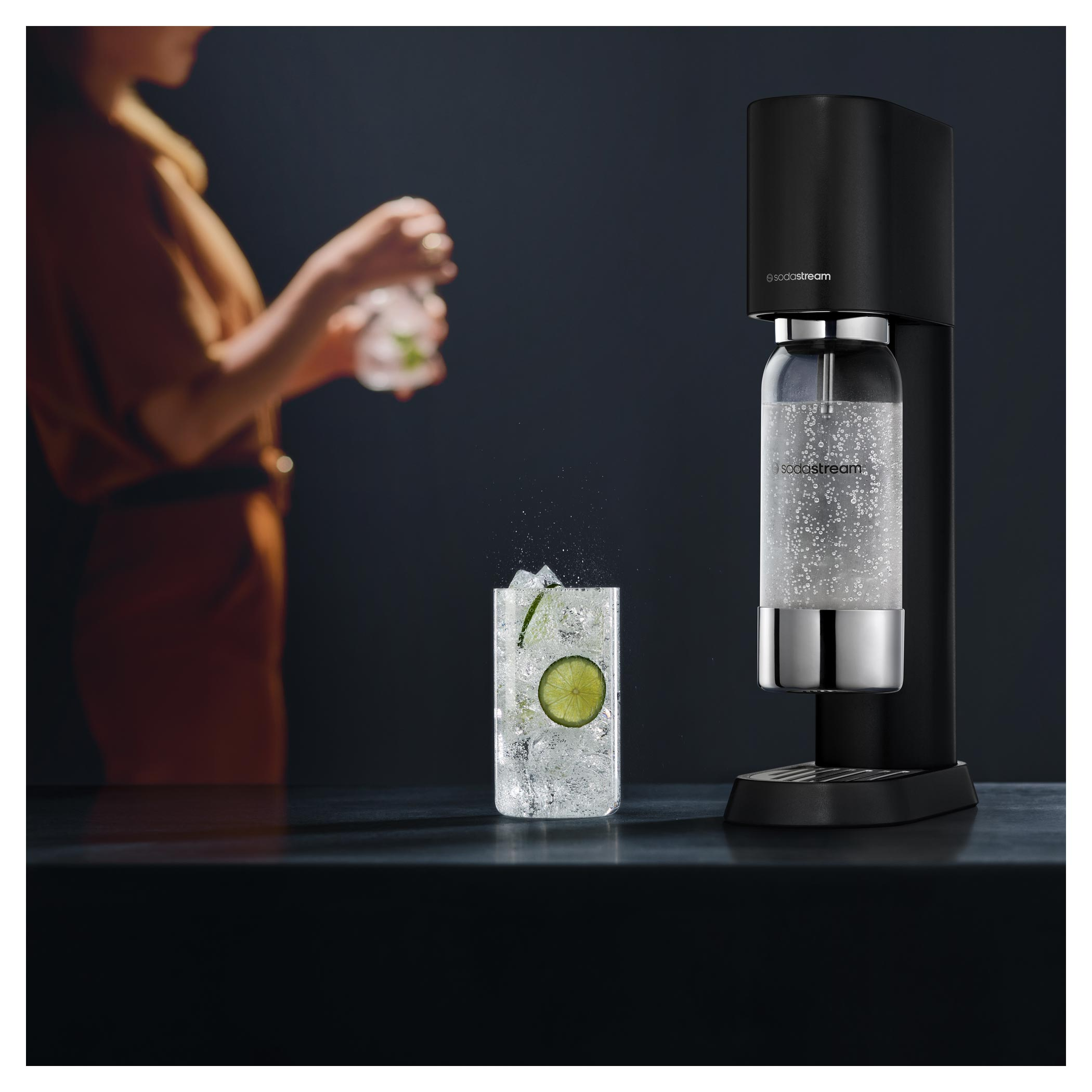 Egy SodaStream gép egy pohár itallal, lime szelettel, jégkockákkal és egy személy, aki italt tart.