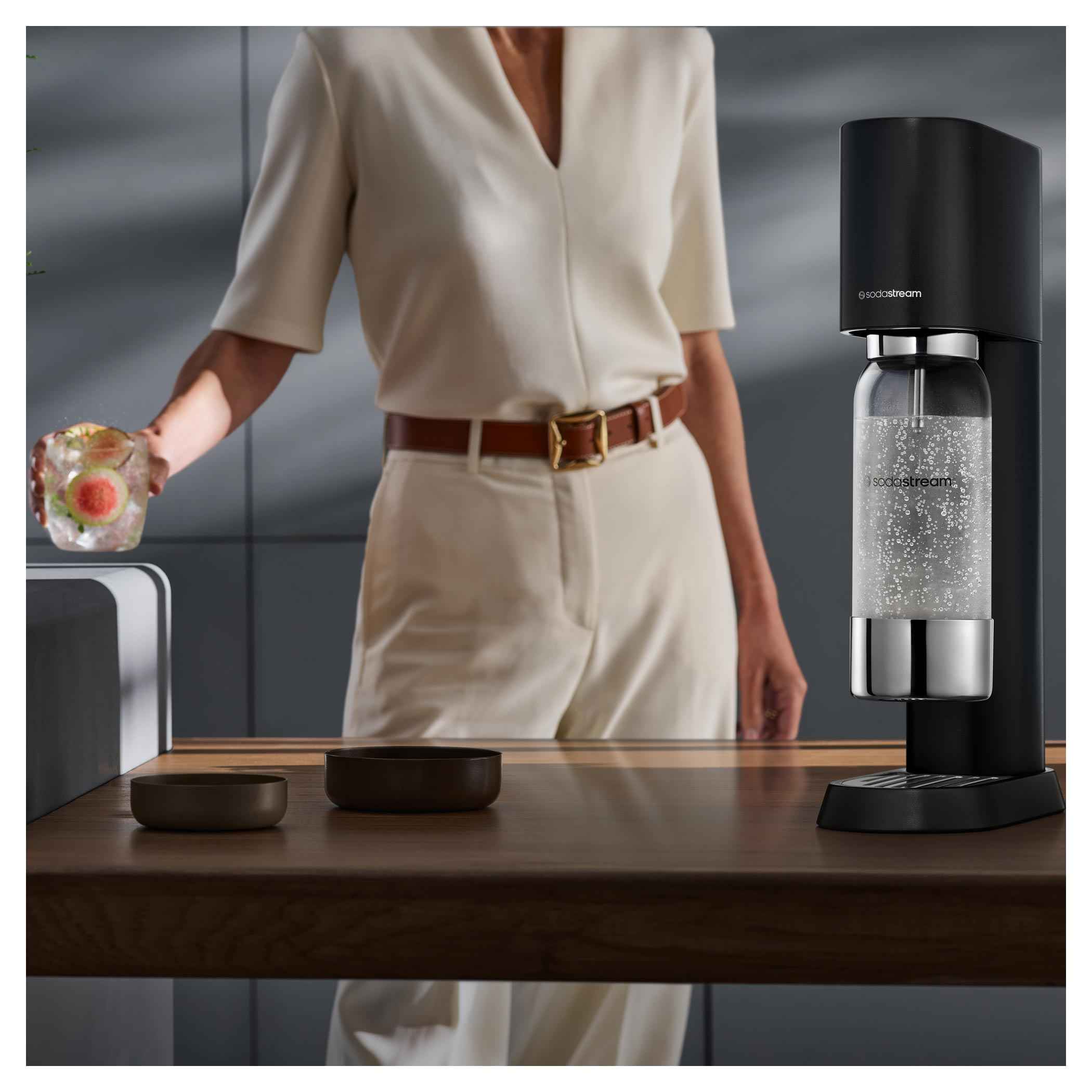Egy személy gyümölcsös koktélt tart a SodaStream közelében. Barna tálak és egy szódakészítő fa felületen.