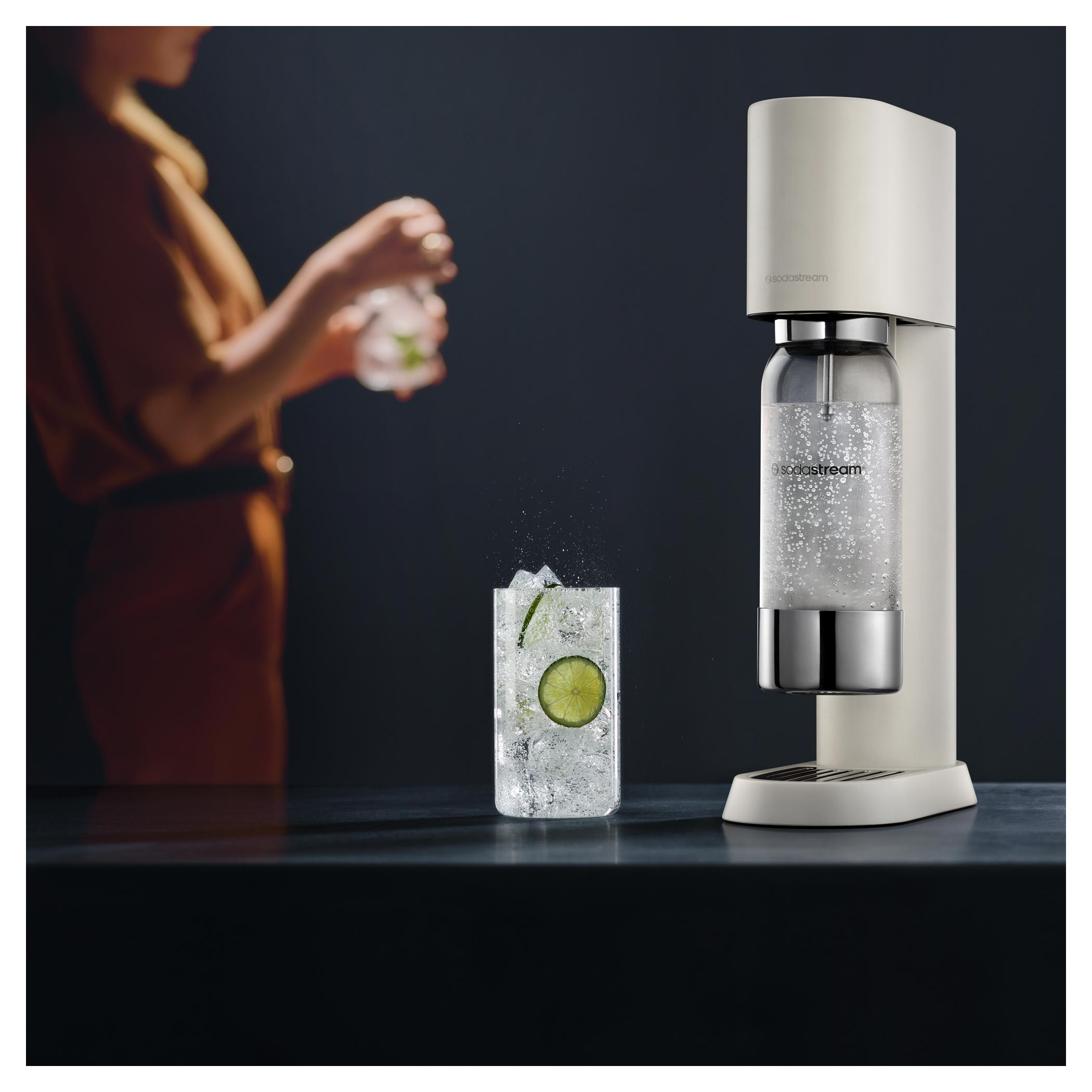 Krémszínű SodaStream, egy pohár jéggel és lime-mal, és egy személy, aki italt készít. Sötét háttér, asztallal.