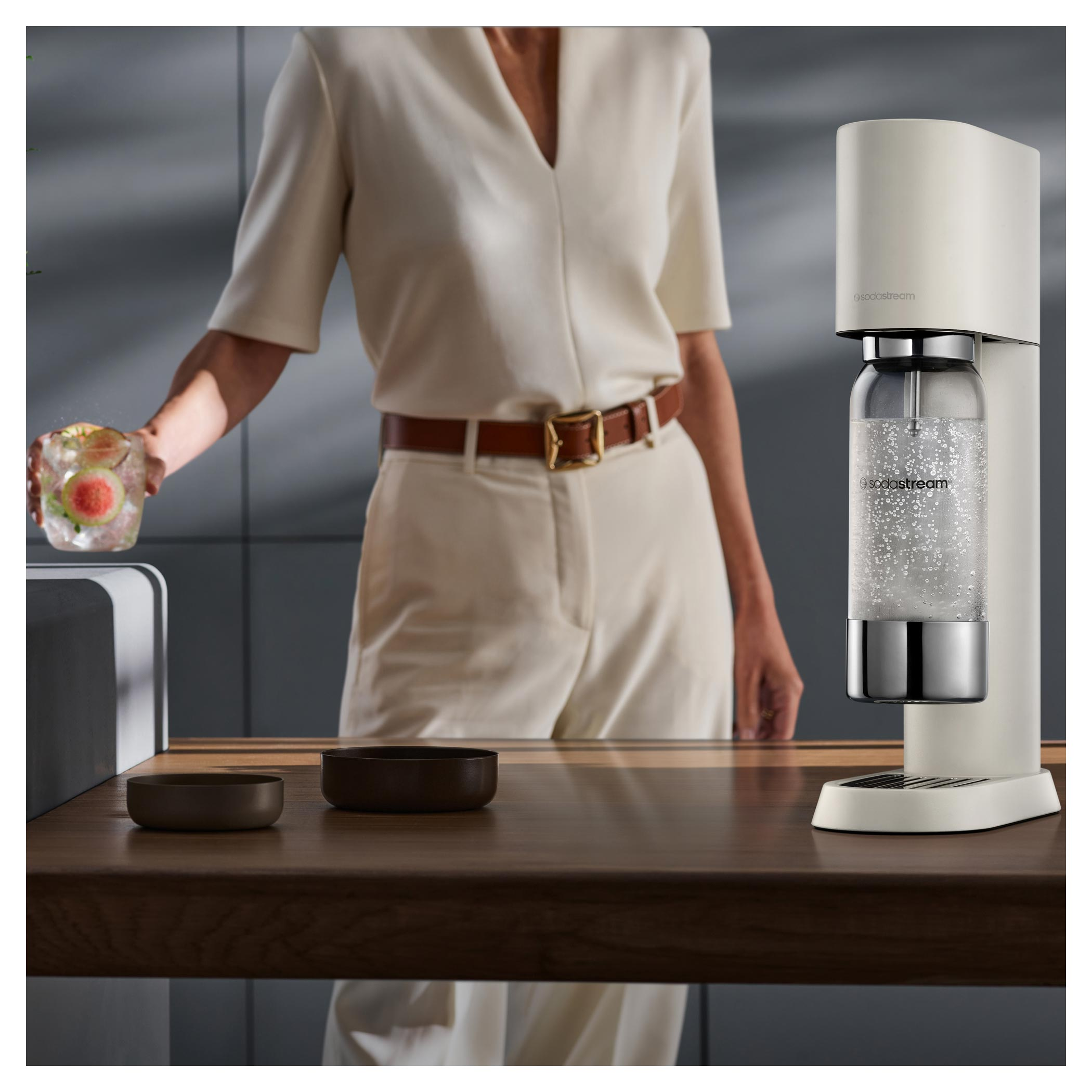 Egy személy gyümölcsös koktélt tart. A SodaStream gép jobb oldalon. Sötét fa asztal tálakkal.