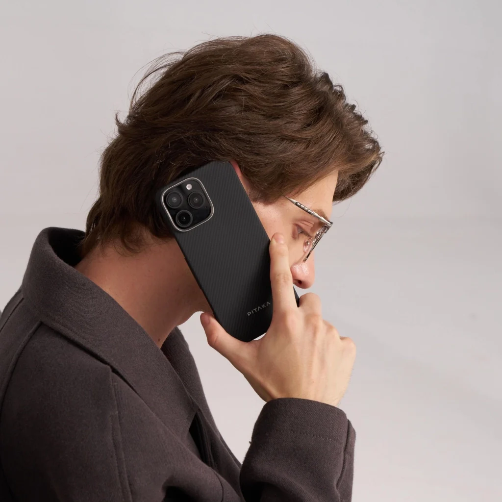 Egy szemüveges férfi egy fekete telefont tart a füléhez. Sötét kabátot visel, és a telefon tokján a PITAKA márkanév látható.