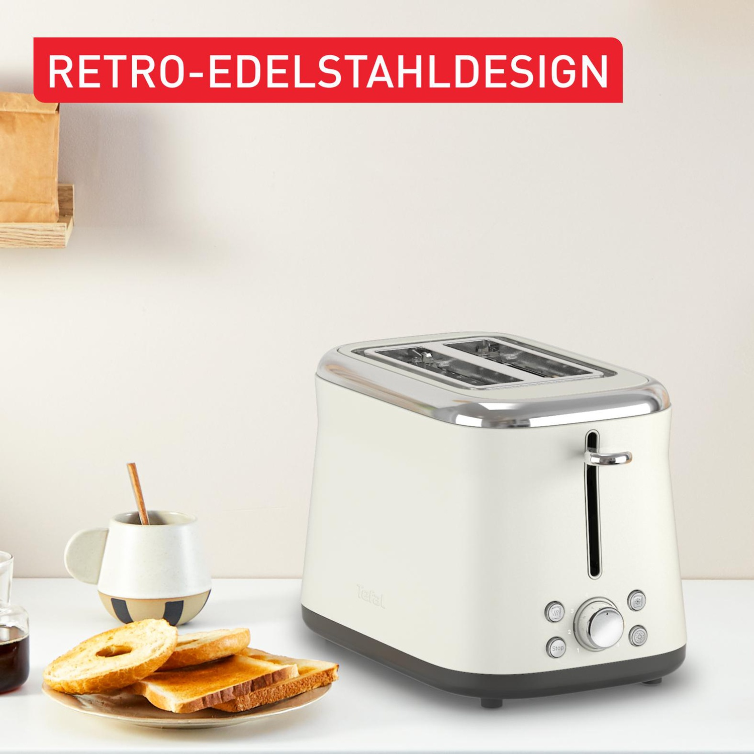 Un grille-pain crème avec des accents chromés est posé sur une surface blanche. Du pain grillé et une tasse sont à côté. Le mot 'Tefal' est visible.