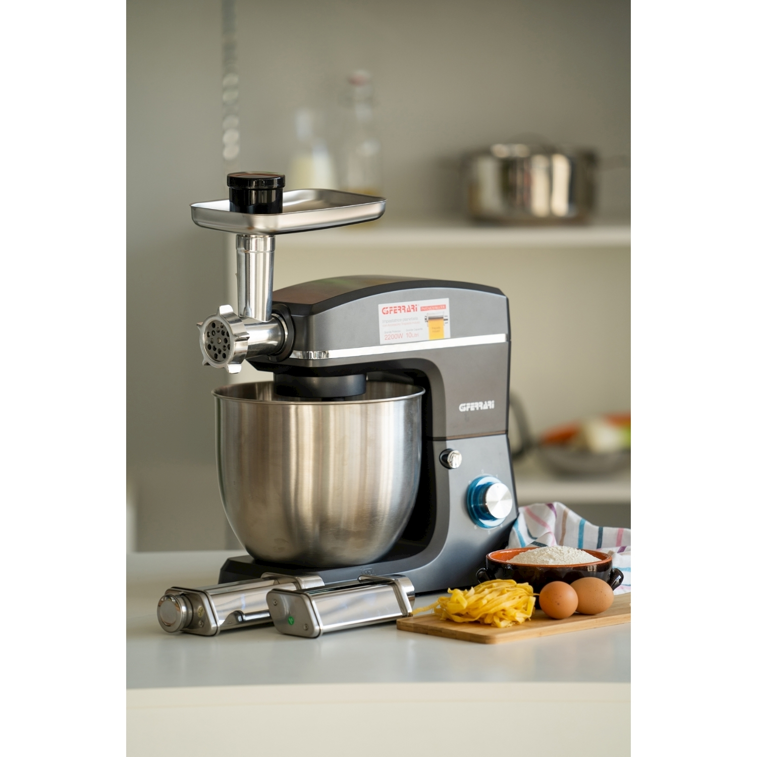 Un robot da cucina grigio con accessori. Pasta, uova e farina su un tagliere di legno. Sfondo: pentole in acciaio inox e una mensola bianca.