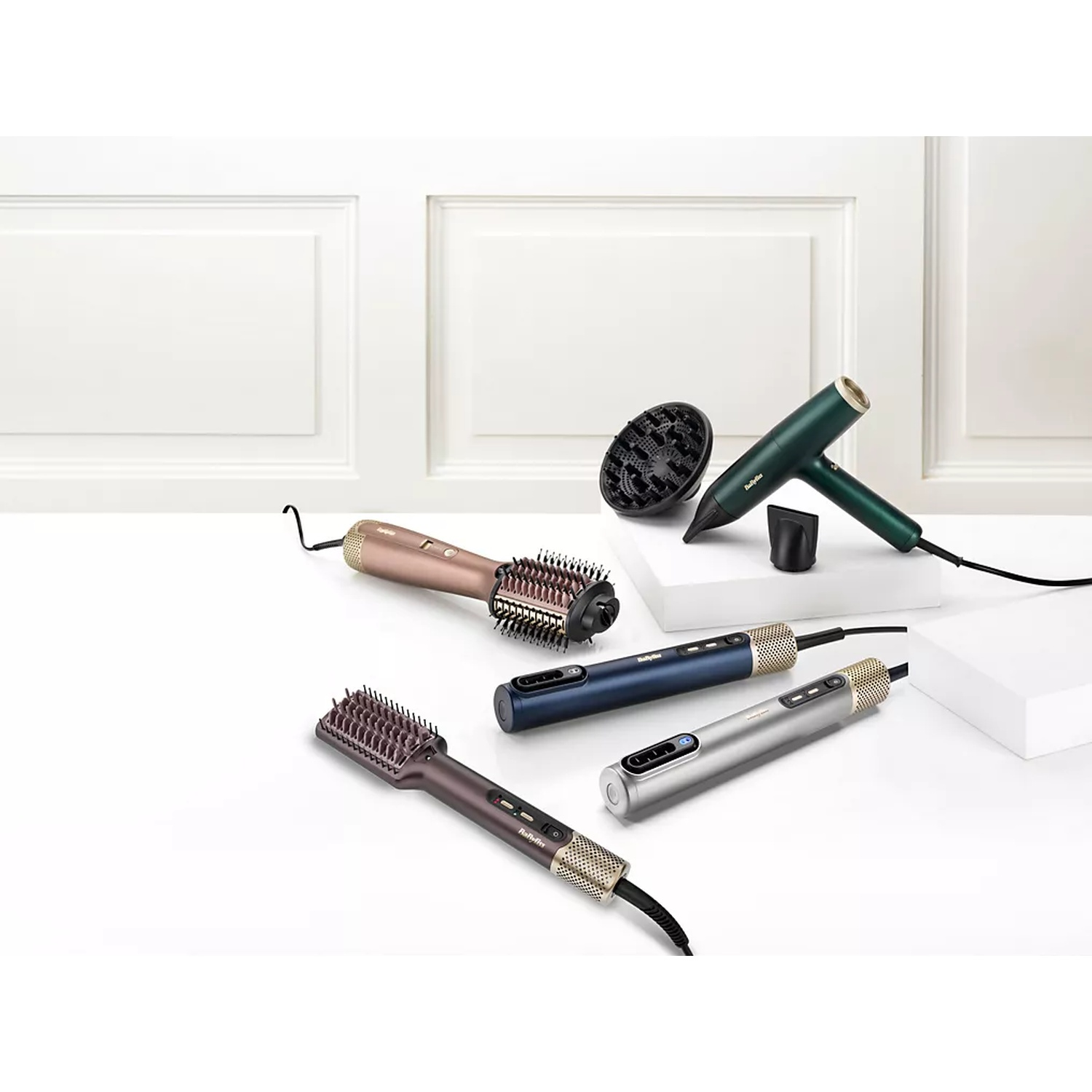 Hair styling tools are arranged on a white surface. Includes hair dryers and brushes in various colors. White wall in the background.