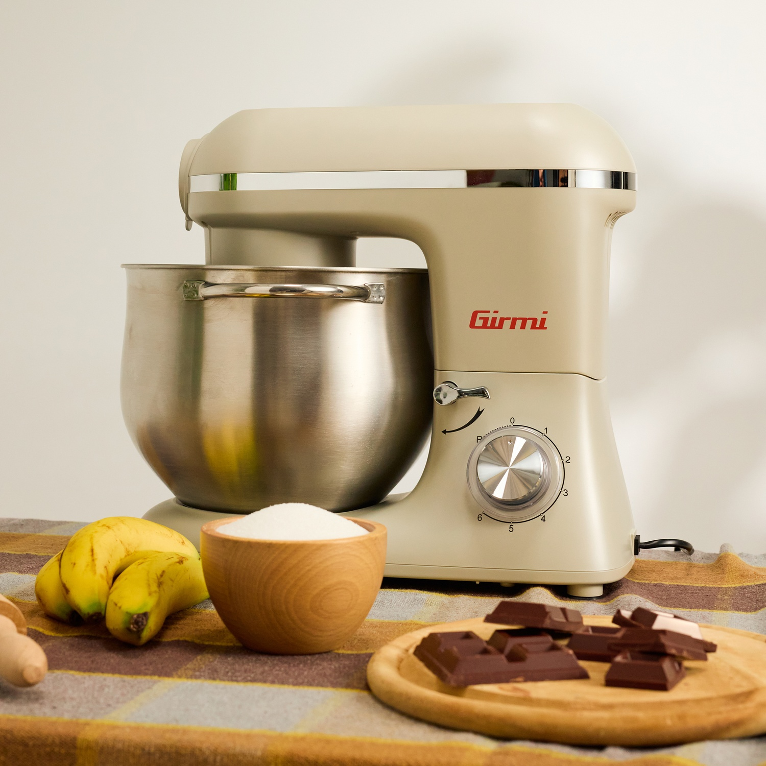 Un'impastatrice beige, una ciotola e ingredienti come banane, zucchero e cioccolato su un panno fantasia, pronti per cuocere.