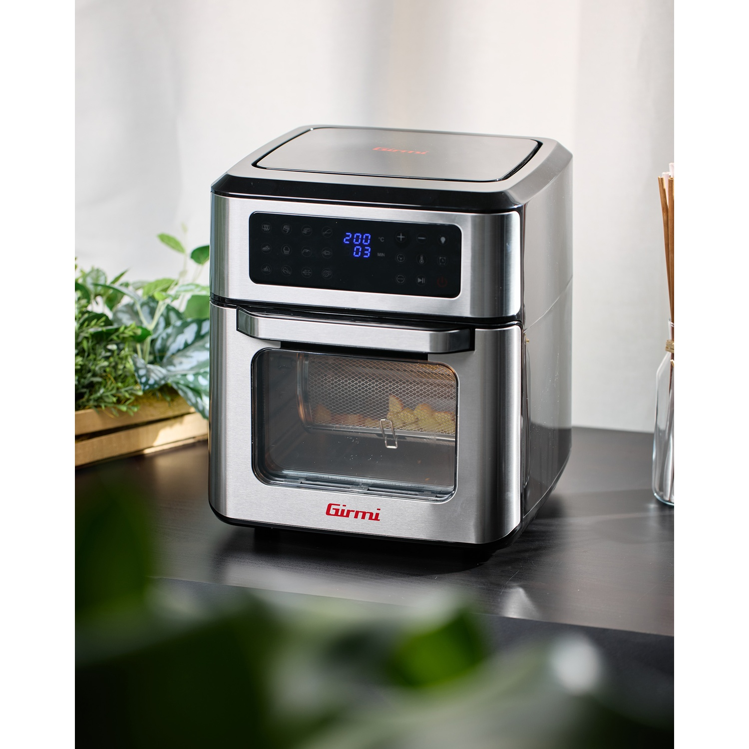 Una friggitrice ad aria in acciaio inossidabile con display digitale poggia su una superficie scura.  All'interno c'è del cibo.  Sono visibili anche piante verdi e un vaso.