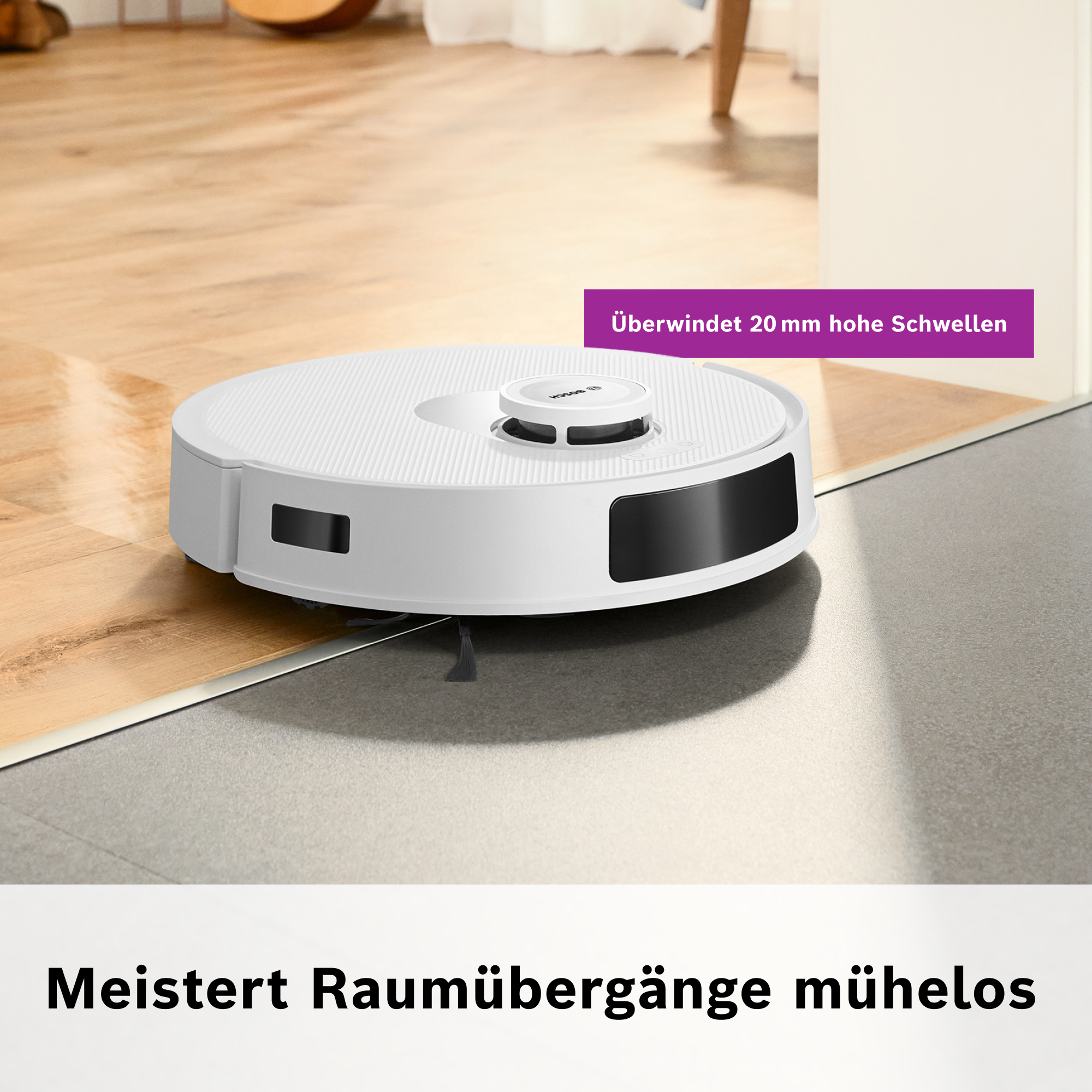Ein weißer Roboter-Staubsauger auf einem Holzboden. Er befindet sich in der Nähe eines Übergangs zu einem grauen Boden.