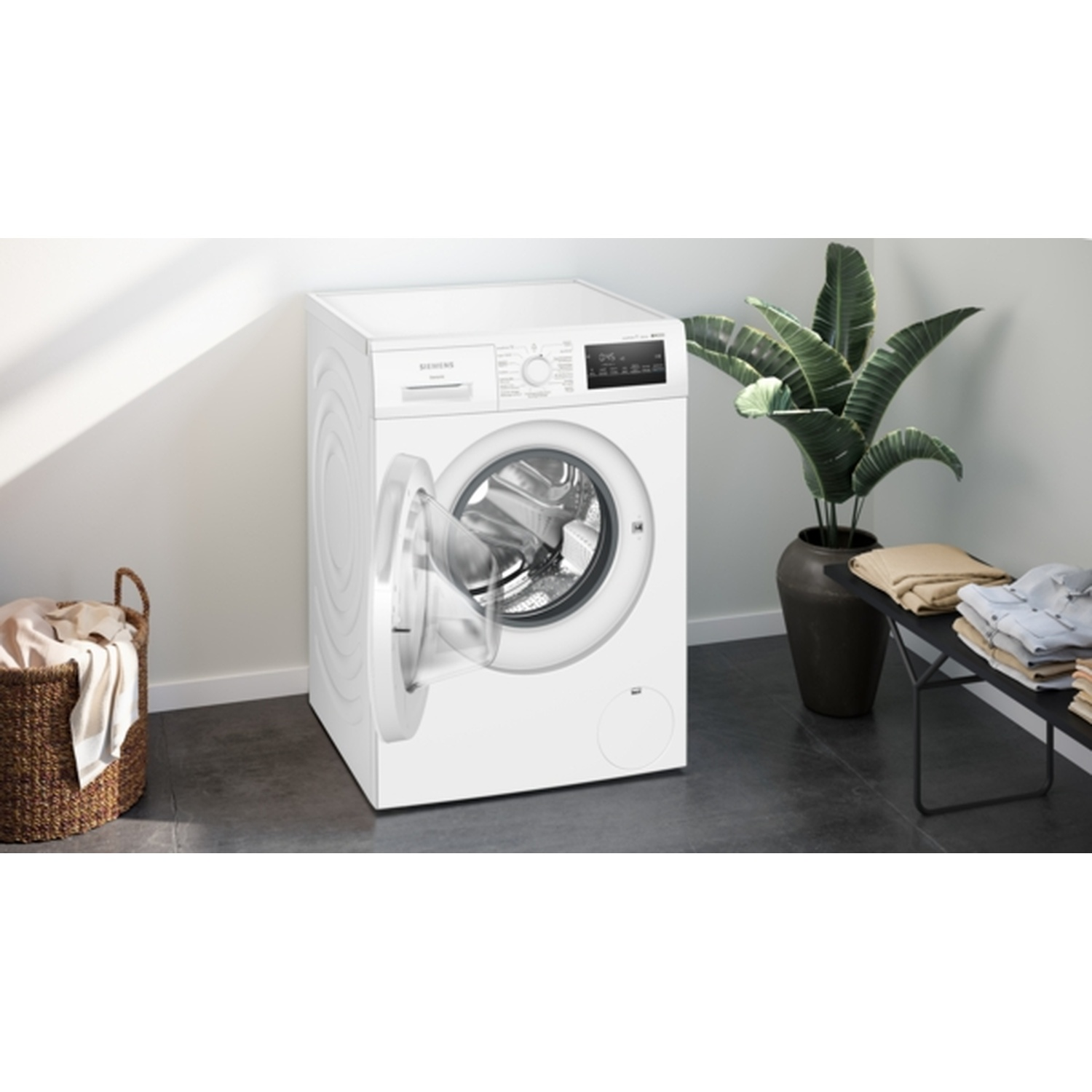 A white washing machine with an open door stands near a laundry basket and a plant. Clothes are folded on a bench.