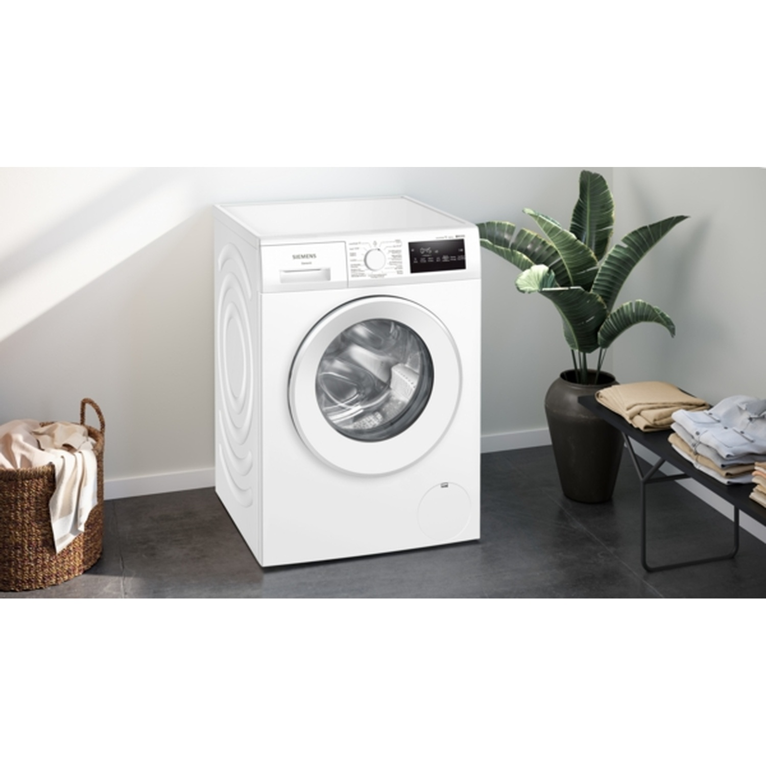 A white washing machine is in a laundry room. A laundry basket on the left, and a plant and folded clothes on the right.