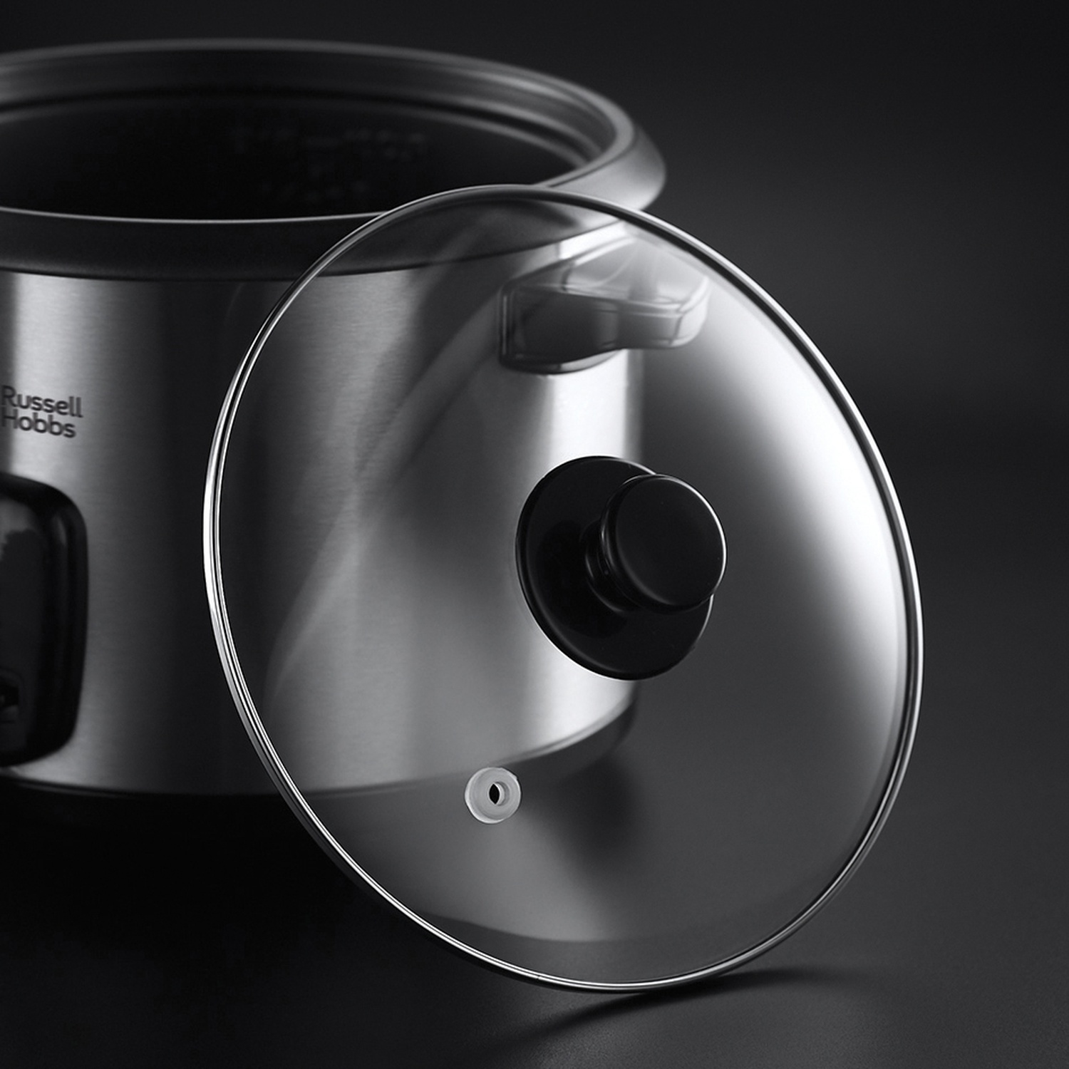 Una cuociriso Russell Hobbs in acciaio inossidabile con coperchio in vetro. Il coperchio ha una maniglia nera. Sfondo nero.