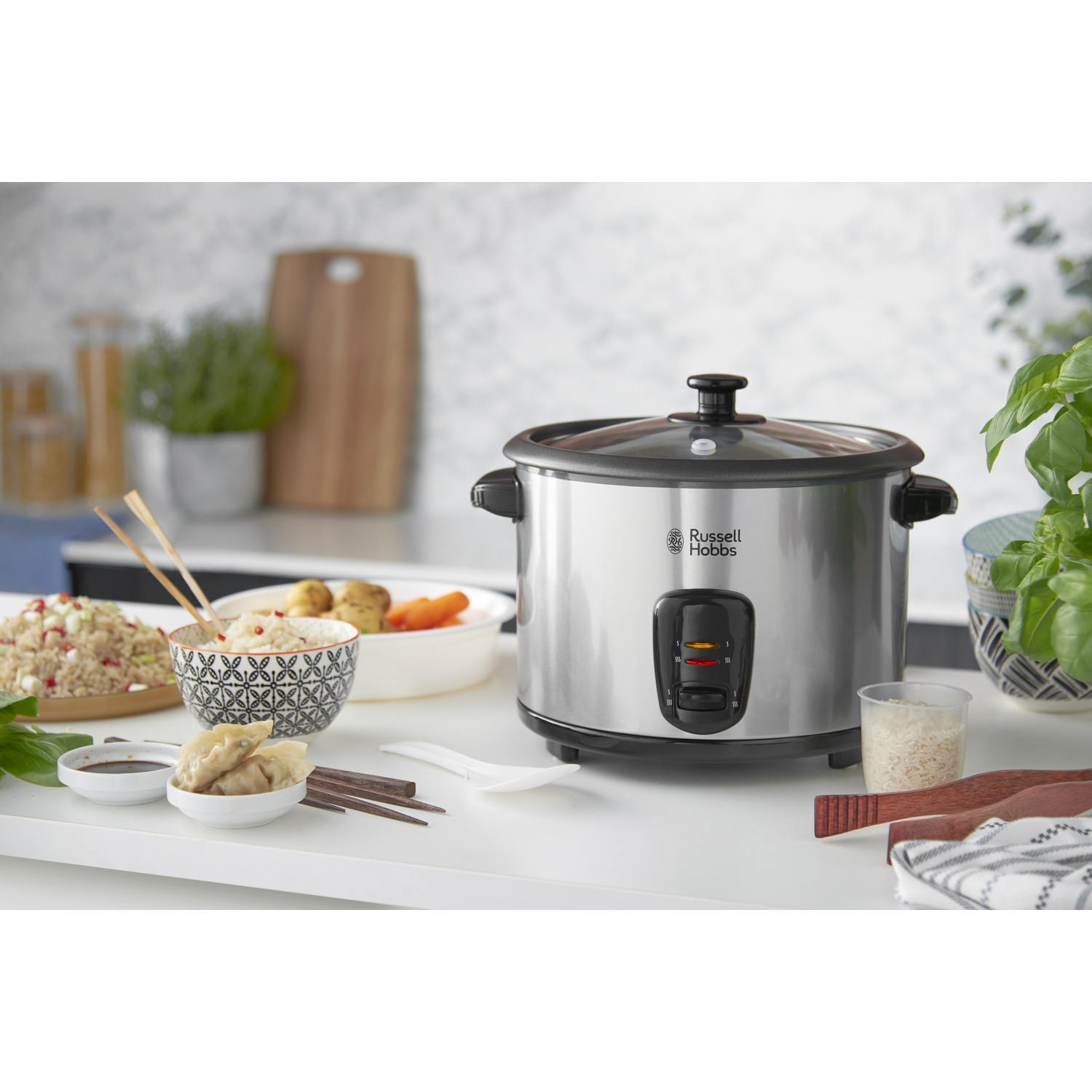 Una cuociriso Russell Hobbs in acciaio inox su una superficie bianca. Ciotole di cibo, bacchette e verdure nella scena.