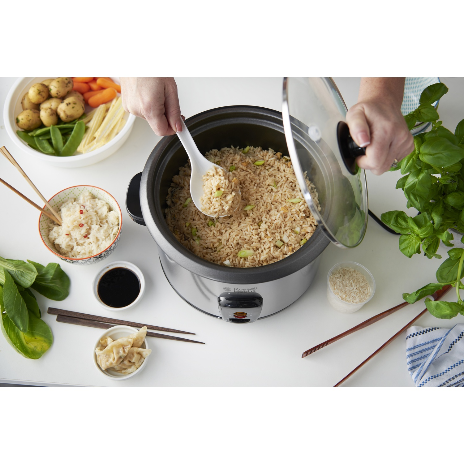 Una persona tira fuori il riso fritto da una cuociriso. Verdure, riso e salsa di soia la circondano. Bacchette e basilico sono vicini.