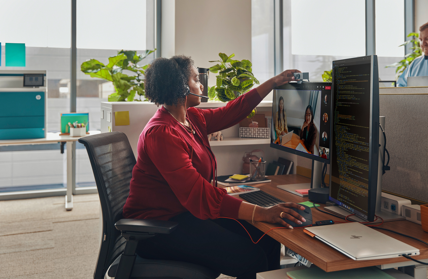 Una mujer con camisa roja está en una videollamada, ajustando la cámara en un monitor. En el escritorio hay un portátil y un teclado.