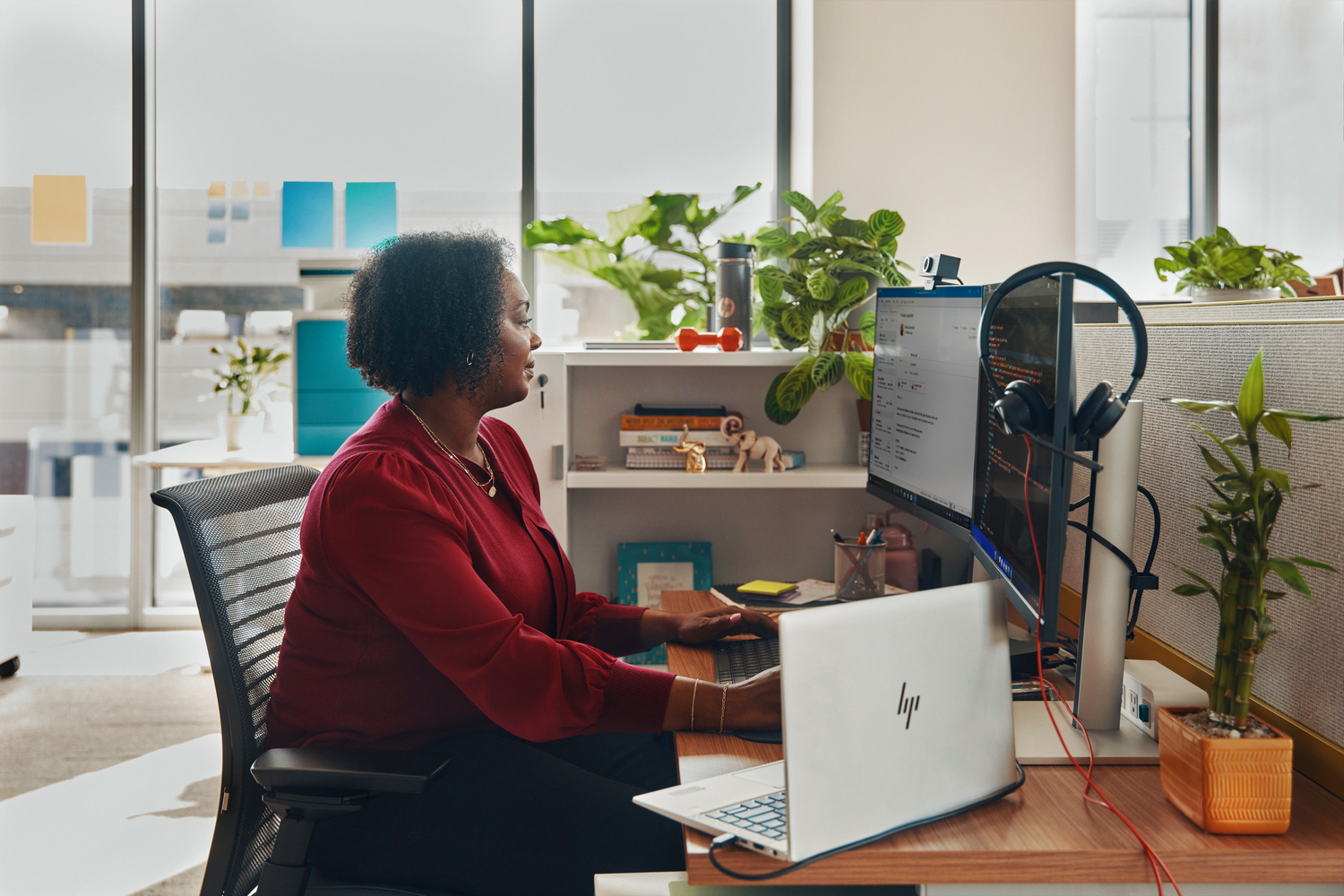 Una mujer con camisa roja trabaja en un escritorio con computadora y portátil. Hay plantas, una ventana y auriculares.
