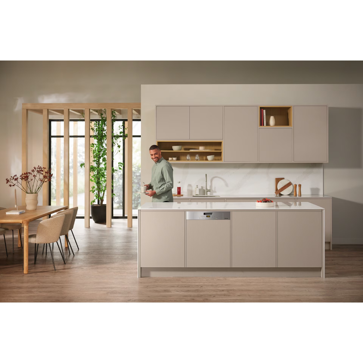 Een man staat lachend in een moderne keuken. Beige kasten, een groot eiland, houten vloer en een eettafel zijn zichtbaar.