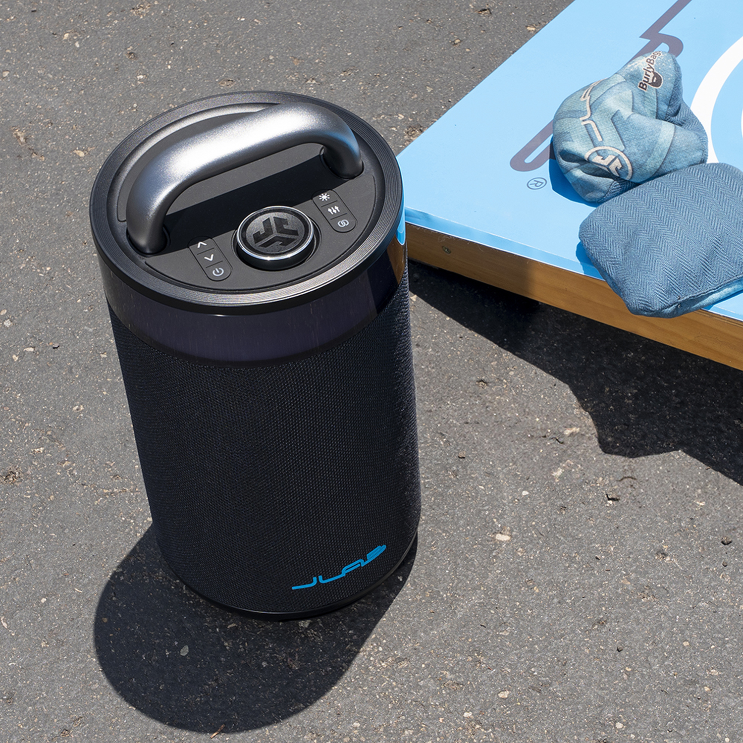 Een zwarte speaker met een handvat staat op asfalt naast een blauw cornholebord met zitzakken.