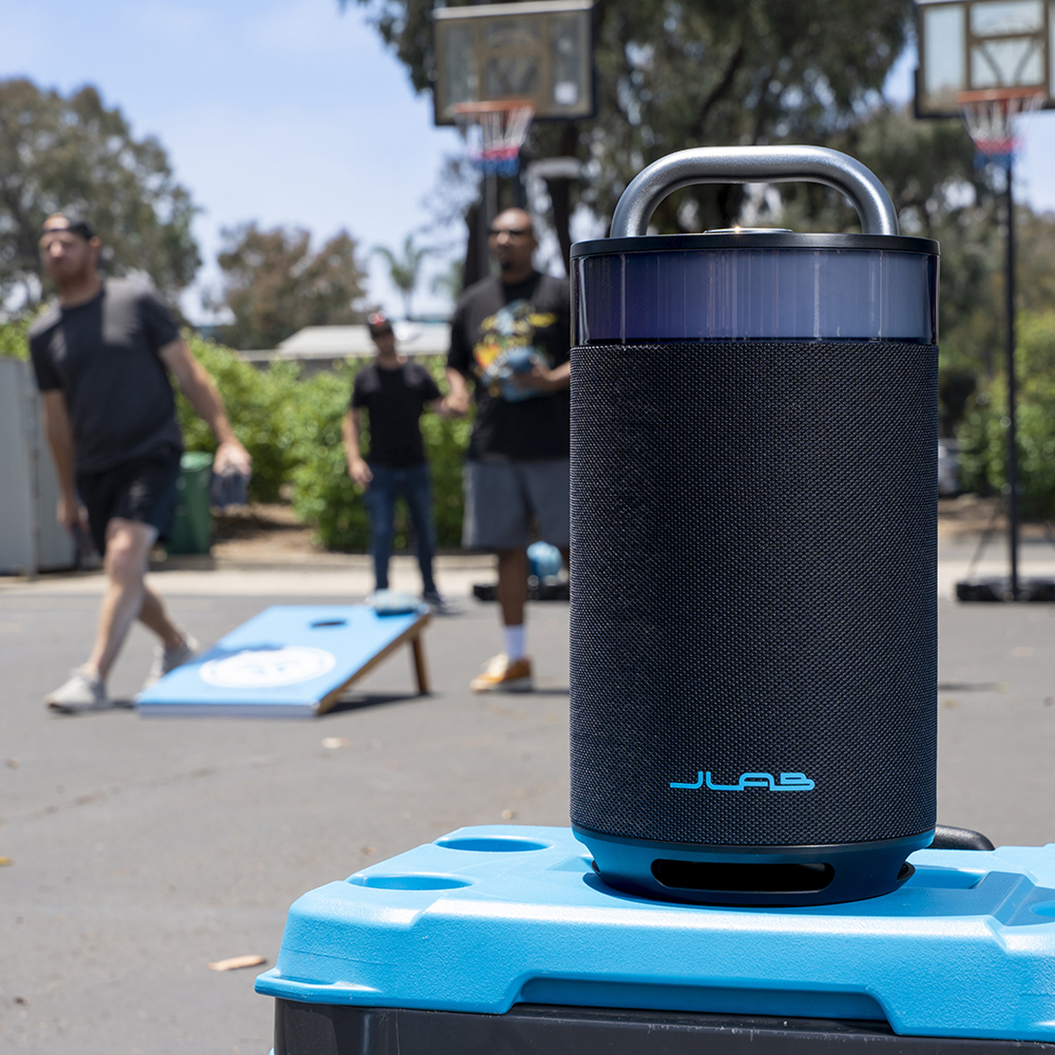 Een zwarte luidspreker staat op een blauwe koeler. Mensen spelen cornhole en basketbal op de achtergrond. De lucht is blauw en zonnig.