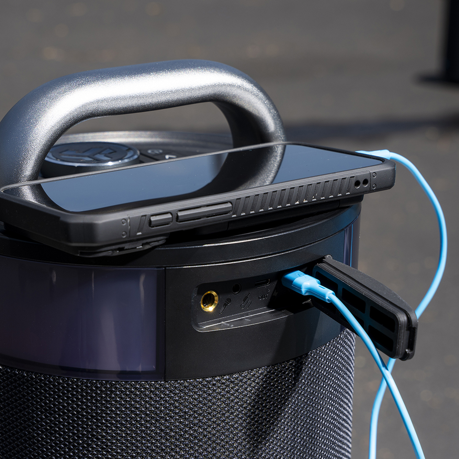 Een zwarte telefoon met hoesje rust op een zwarte luidspreker. Een blauwe kabel verbindt de telefoon en de luidspreker. De luidspreker heeft een handvat.