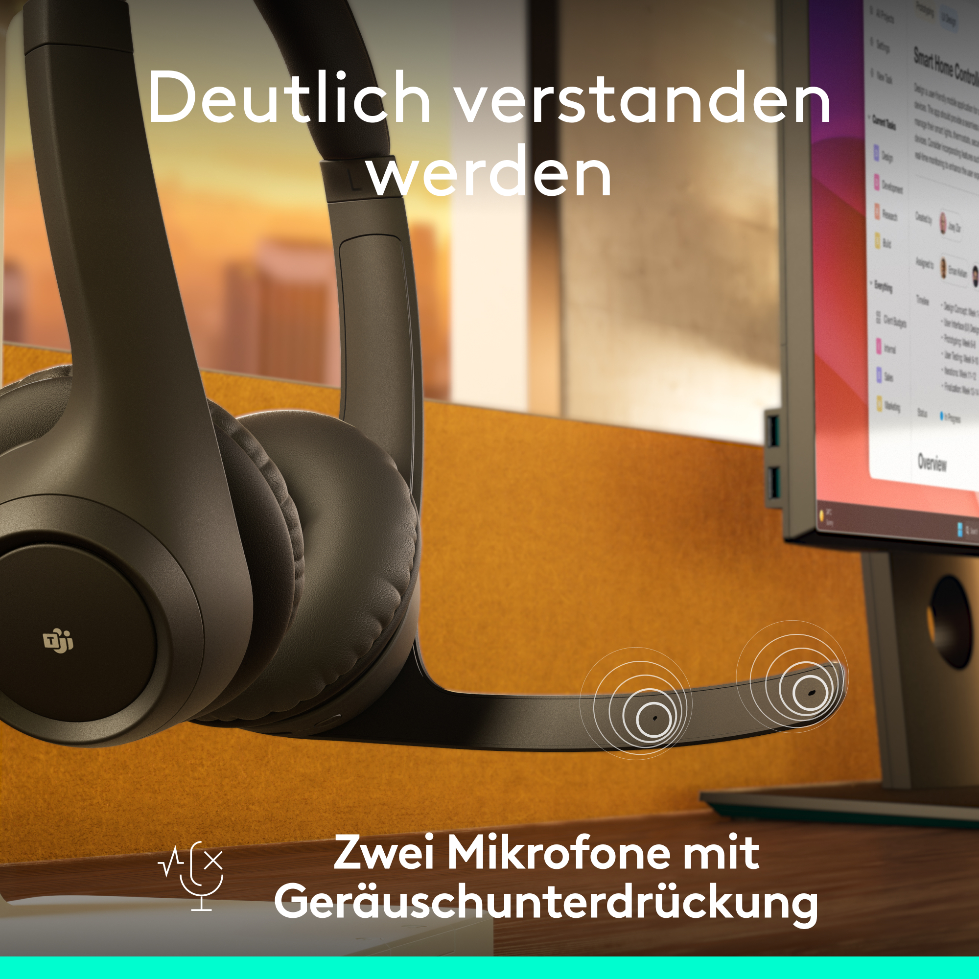 Kopfhörer mit zwei Mikrofonen. Im Hintergrund ein Computerbildschirm. Die Kopfhörer sind schwarz und auf einem Holztisch platziert.