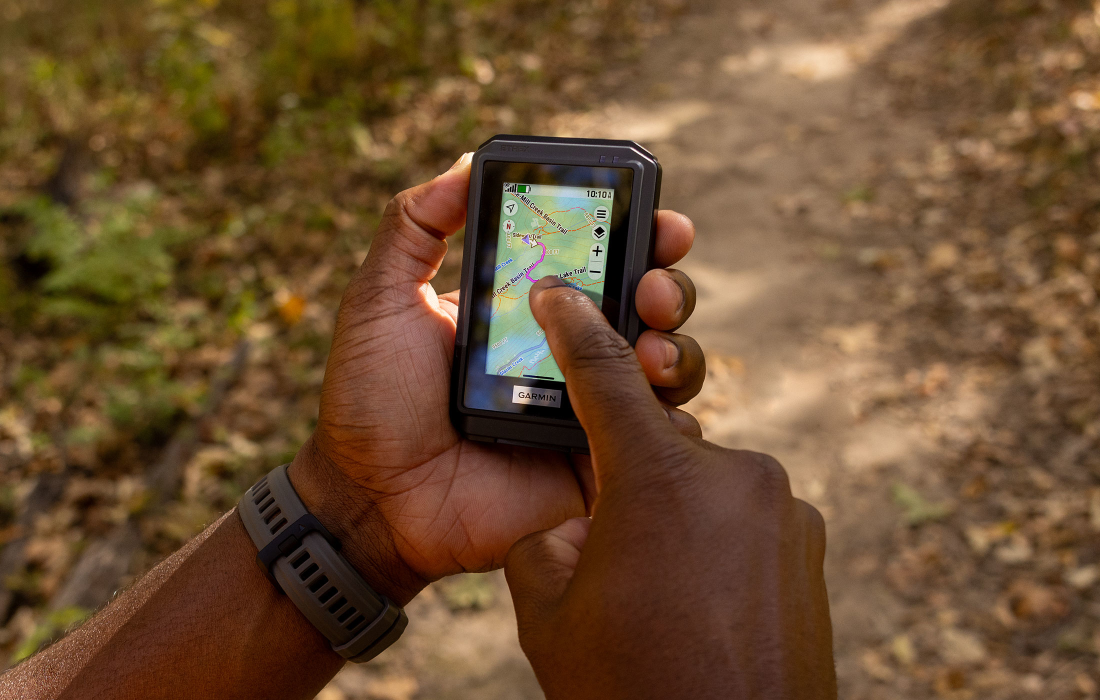 Eine Person hält ein GPS-Gerät und zeigt auf eine Karte. Der Hintergrund ist eine verschwommene Ansicht eines Weges in einem Wald.