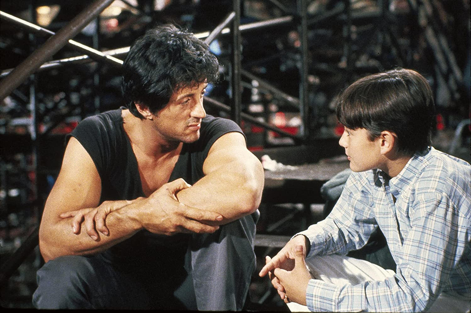 Sylvester Stallone und ein Junge sitzen sich gegenüber. Stallone trägt ein schwarzes Hemd. Der Junge trägt ein kariertes Hemd. Metallgerüst im Hintergrund.