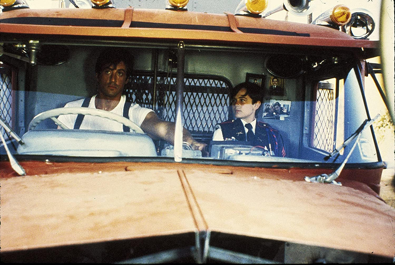 Zwei Männer in einem Oldtimer-Truck. Einer fährt, trägt weißes Hemd und Hosenträger. Der andere in Uniform. Der Truck ist rot.