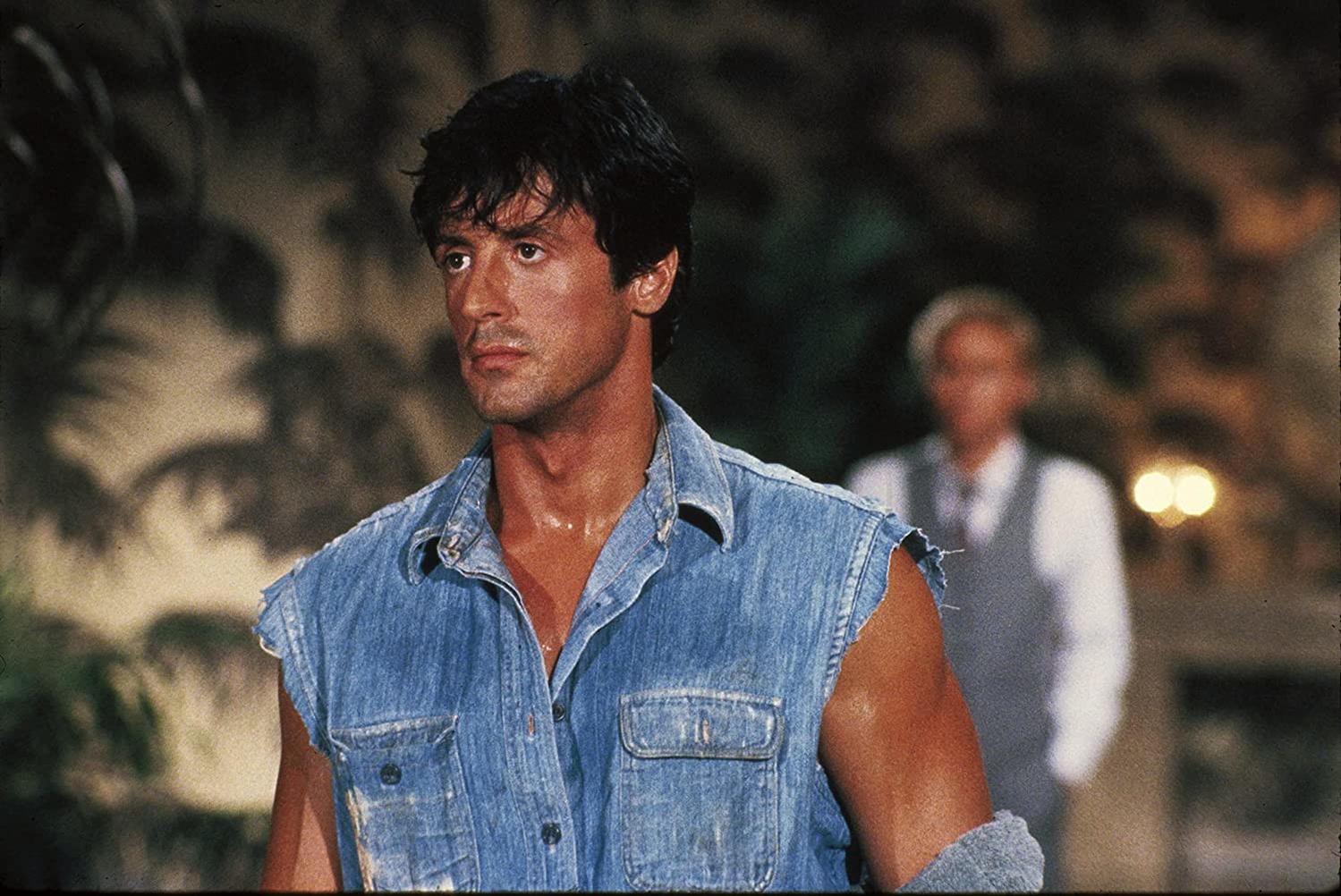 Sylvester Stallone in einer Jeansweste blickt nach links. Ein Mann im Anzug ist im Hintergrund verschwommen. Tropisches Laub.