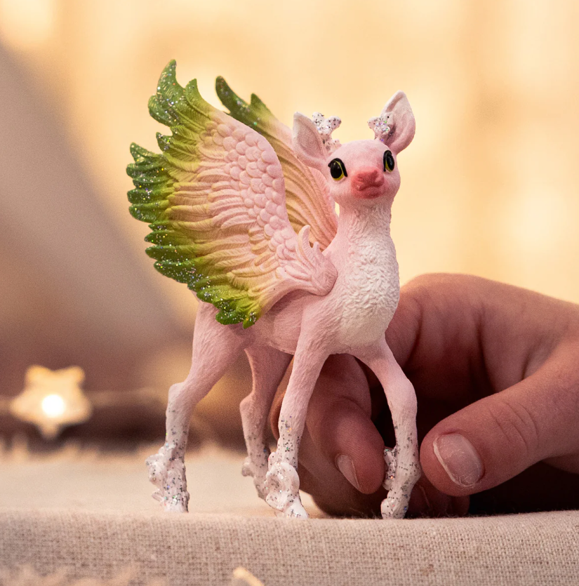 Eine rosa geflügelte Kreatur-Figur mit grünen und glitzernden Flügeln wird von einer Hand gehalten. Es hat Glitzer an den Füßen. Weicher Hintergrund.
