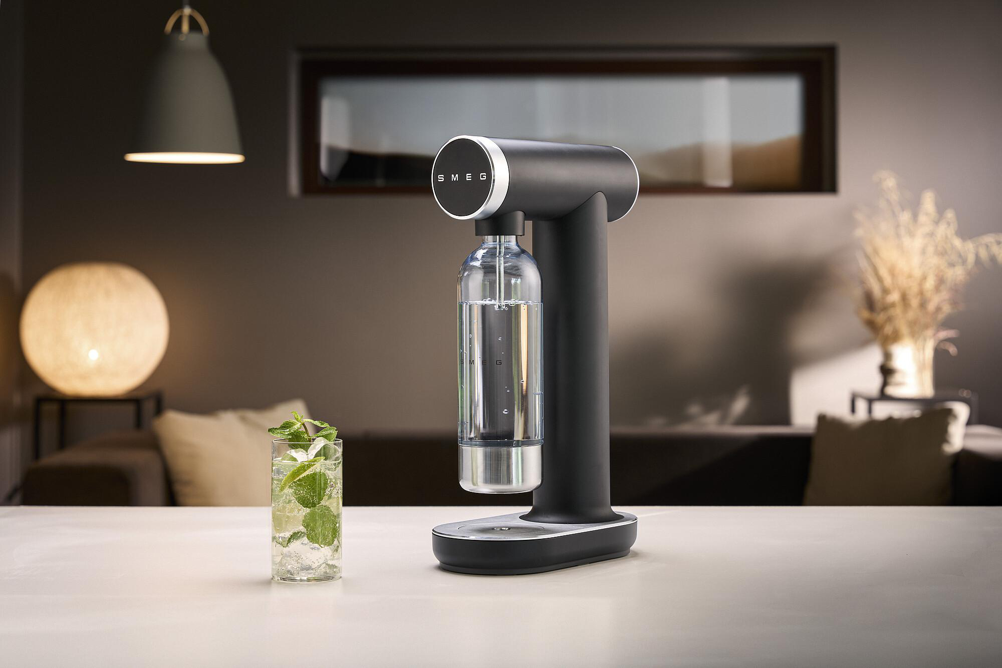 Ein schwarzer SMEG-Sodabereiter mit einem Glas Mojito. Im Hintergrund eine Lampe und ein Fenster.