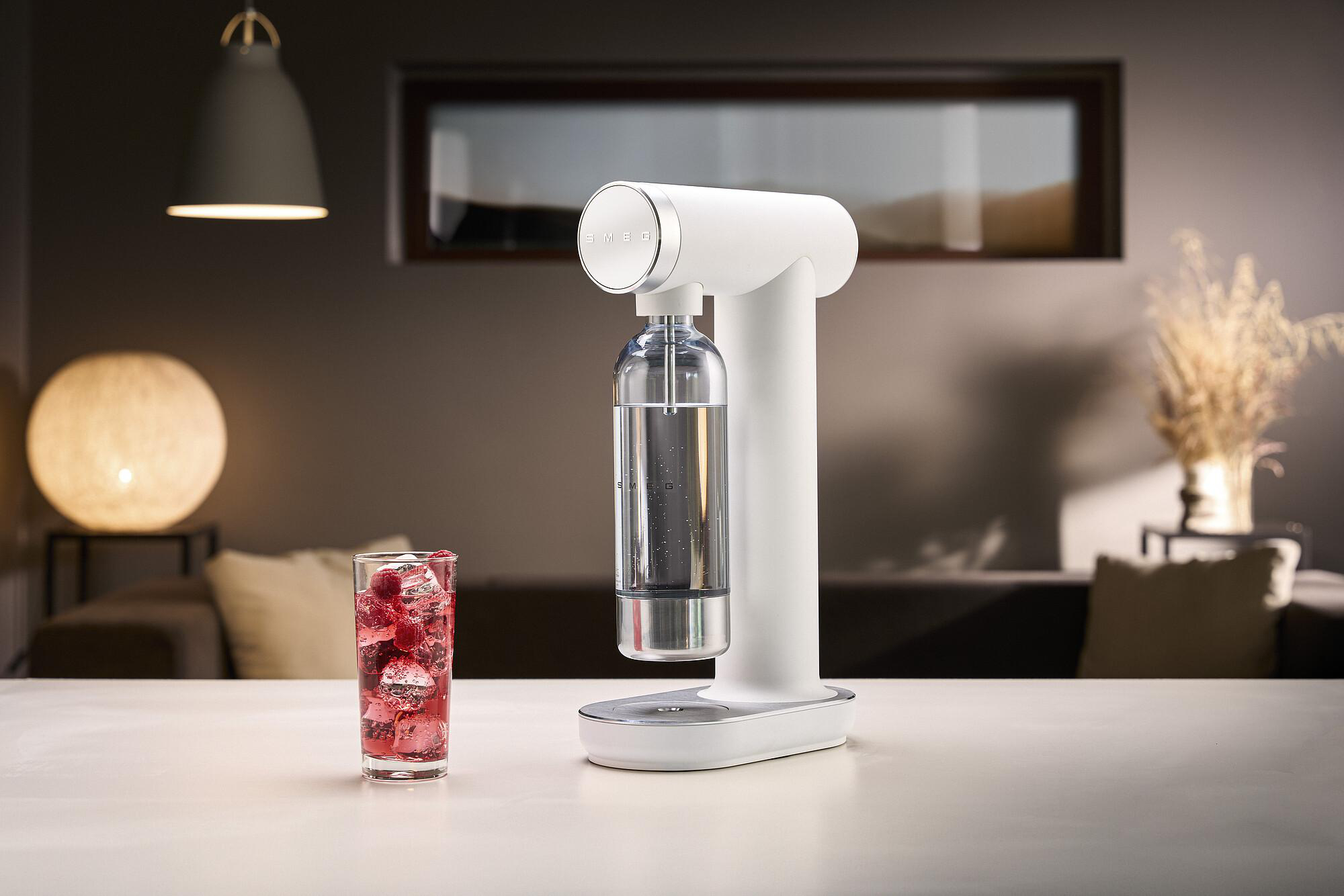 Ein weißer Sodabereiter mit einem Glas rotem Getränk mit Eis. Eine Lampe und ein Fenster sind im Hintergrund.