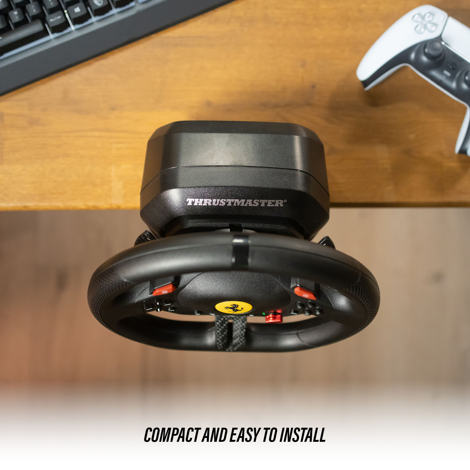 A black Thrustmaster racing wheel, with red buttons, on a wooden desk with a keyboard and a white game controller.