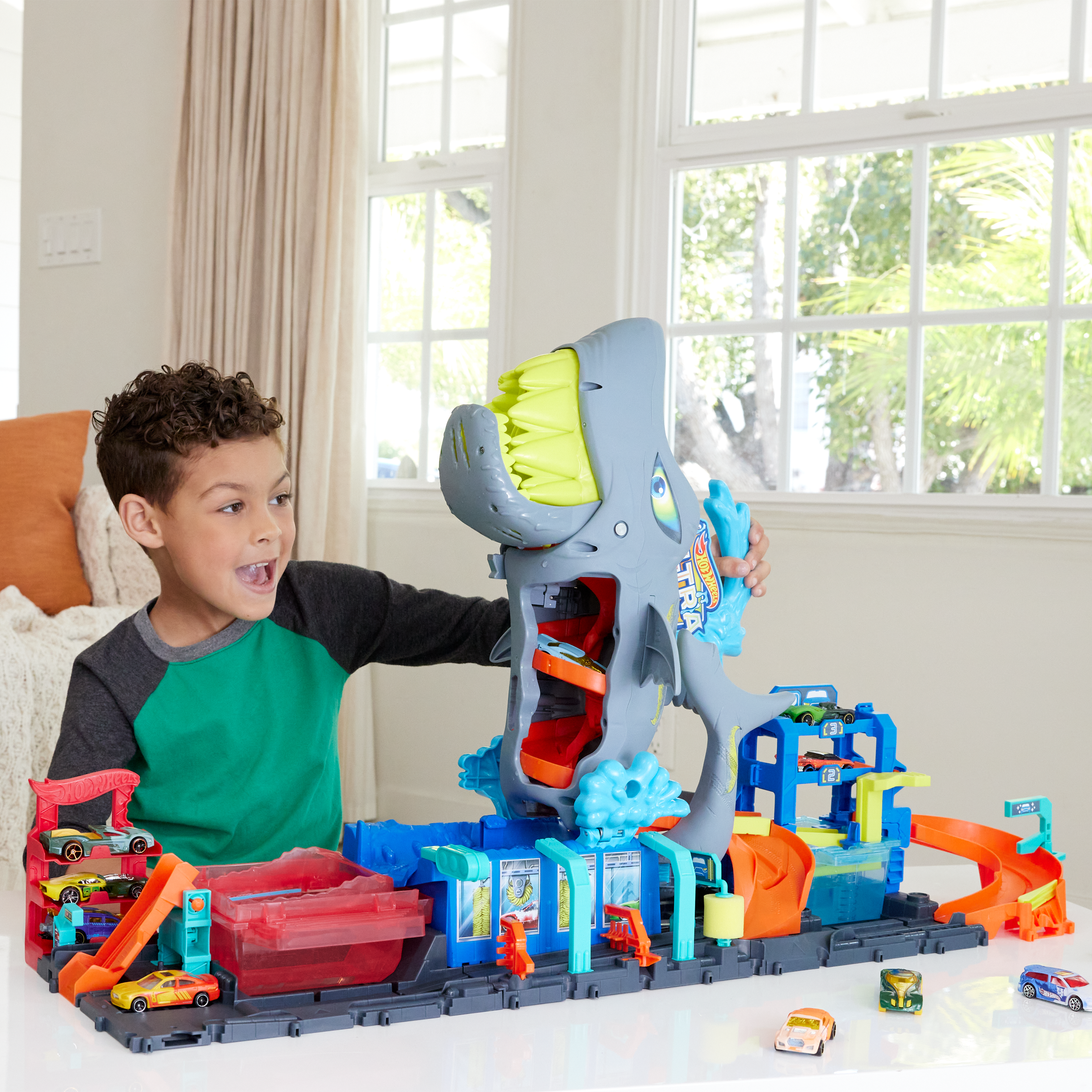 Ein Junge spielt mit einem Hot Wheels Shark Wash Set. Das Set hat einen Haifischkopf, Rampen und bunte Autos. Der Junge lächelt.