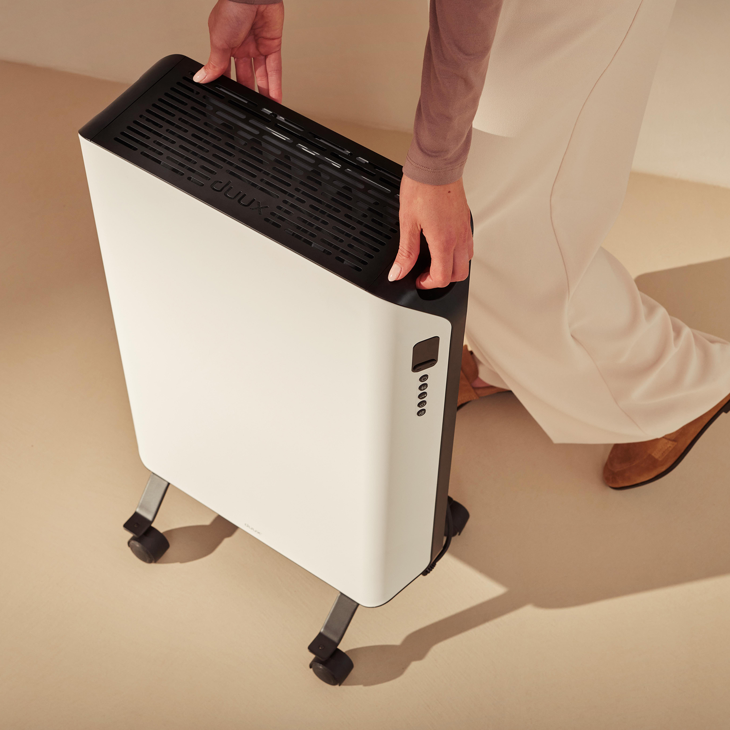 Een persoon houdt een wit en zwart verwarmingstoestel vast. Het toestel staat op wielen. De persoon draagt een beige broek en bruine schoenen.