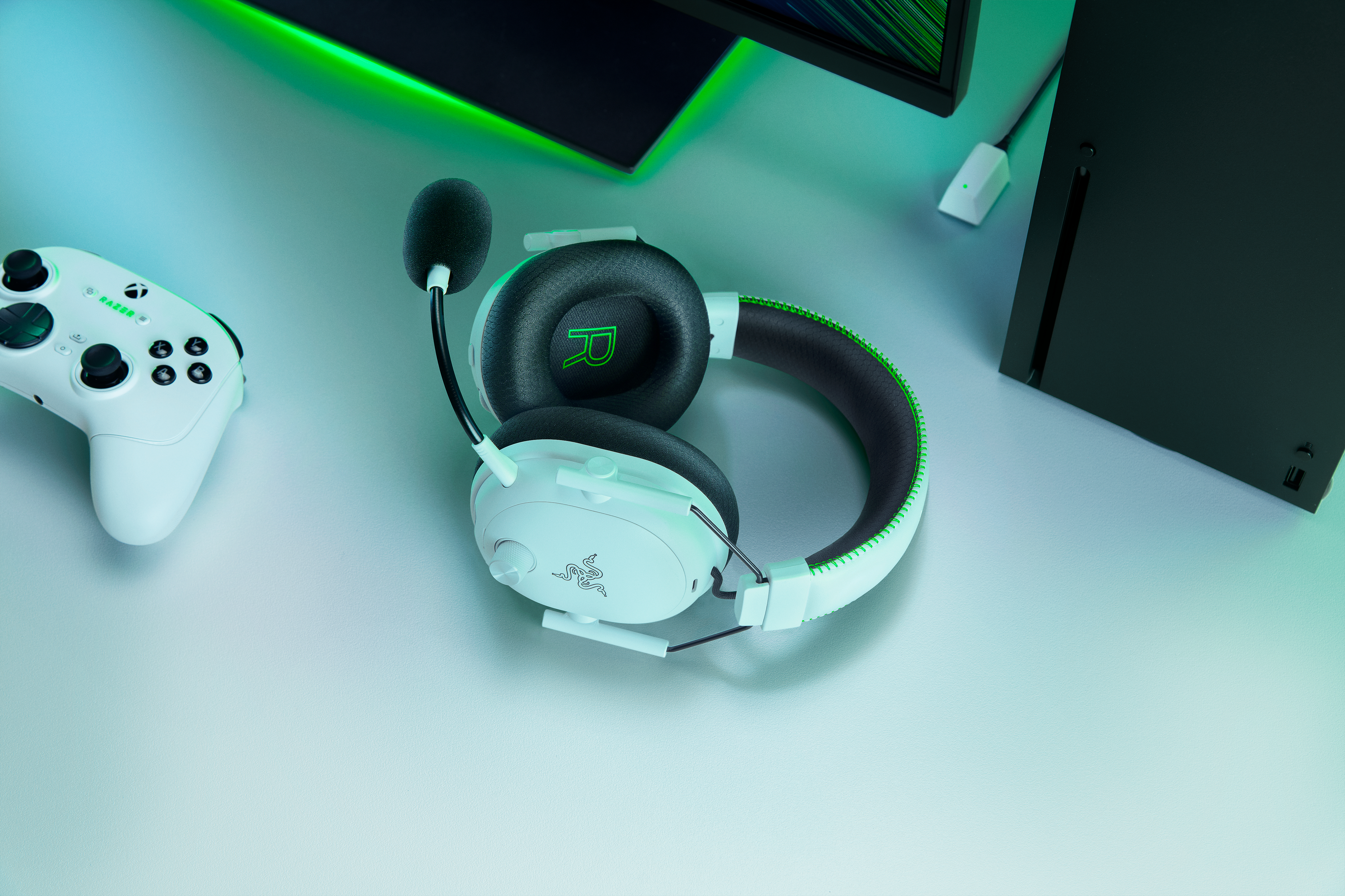 Weißes Headset mit Mikrofon, Controller, schwarze Konsole und Monitor. Razer-Logo vorhanden. Grüne und schwarze Akzente.