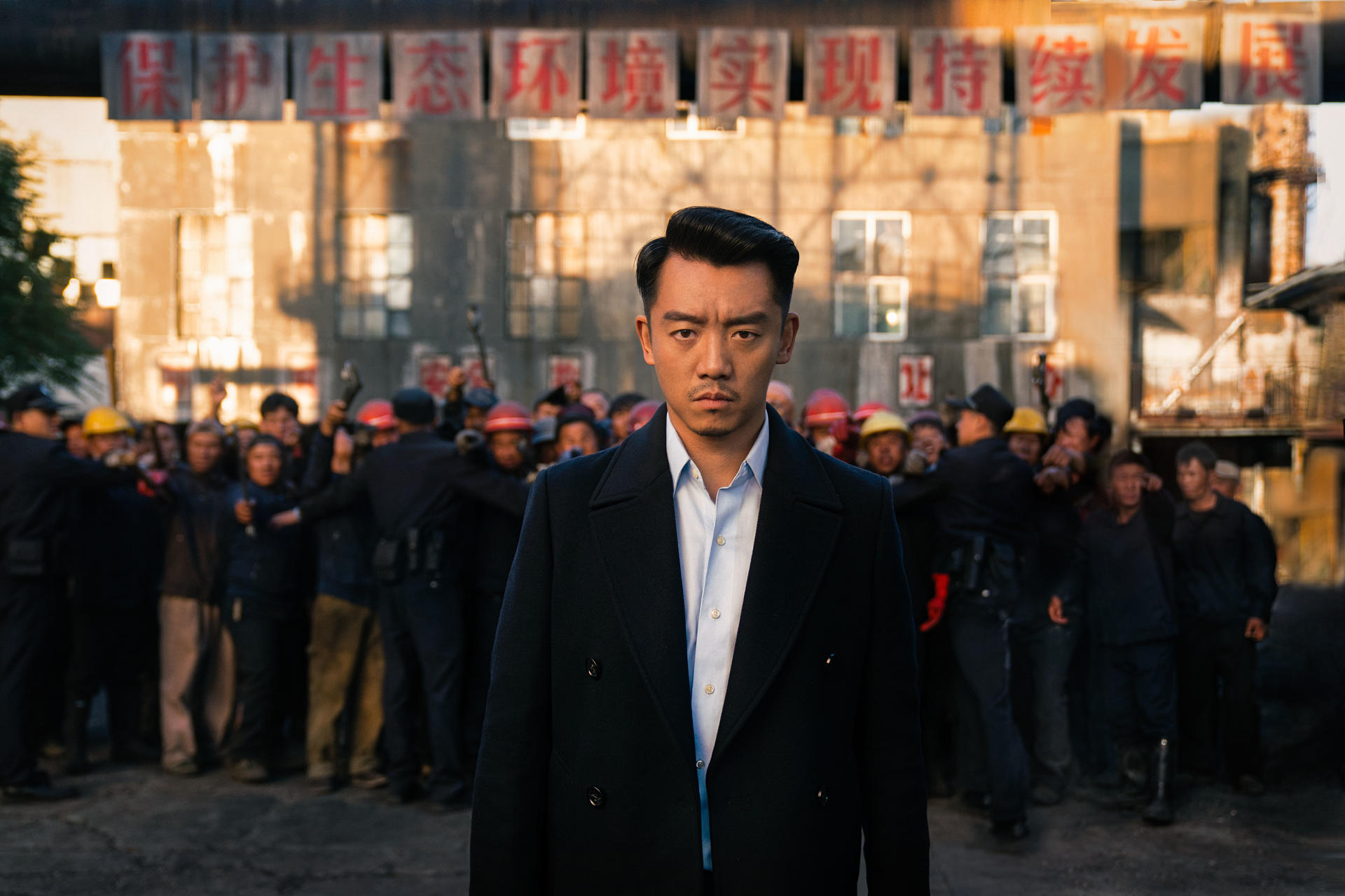 Ein Mann in einem marineblauen Mantel steht vor einer Menschenmenge. Im Hintergrund chinesische Schriftzeichen. Dahinter tragen Menschen Helme und Uniformen.