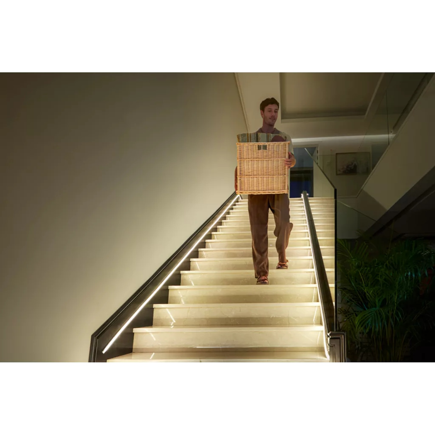 Een man draagt een mand naar beneden op een verlichte trap. Hij draagt een bruine broek en een lichte top. De achtergrond toont een modern interieur.