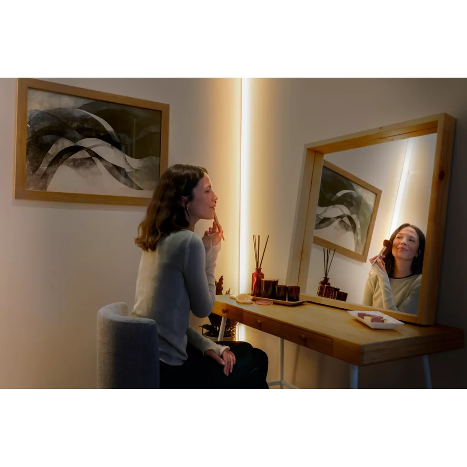 Een vrouw brengt make-up aan bij een kaptafel. Een spiegel weerspiegelt haar beeld. Een schilderij hangt aan de muur en er staan ​​schoonheidsproducten op tafel.