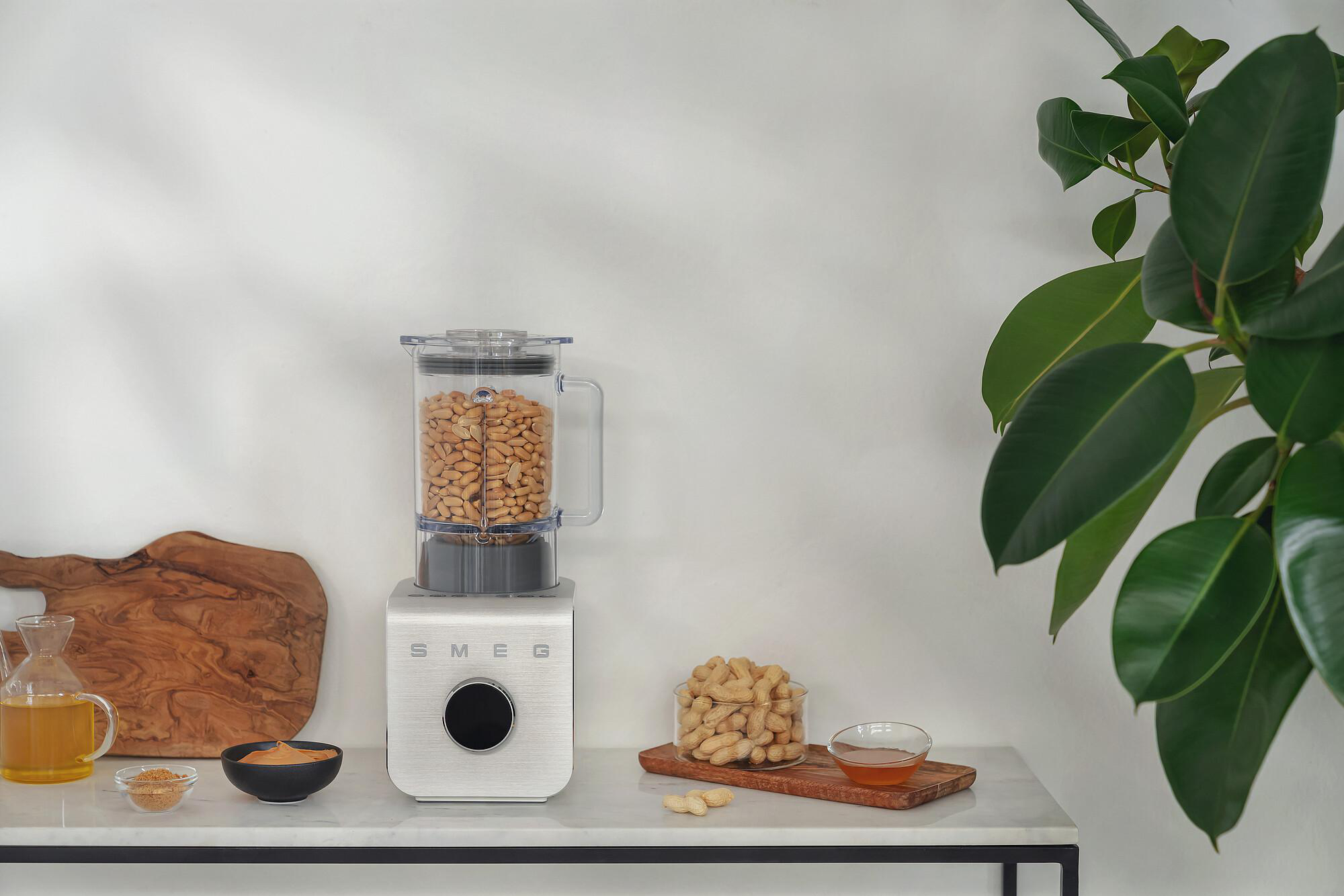 Ein Mixer mit Erdnüssen, umgeben von Zutaten und einer Pflanze. Eine Glas mit Erdnüssen und Honig stehen auf einem Tisch.