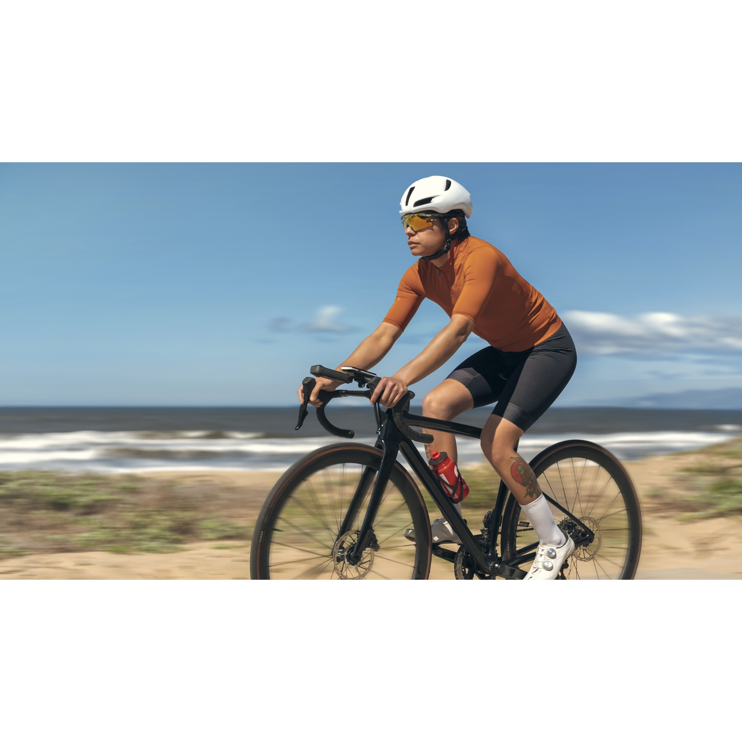 Cycliste sur vélo noir roule le long de la plage. Chemise orange, casque blanc et lunettes de soleil. Océan et ciel bleu en arrière-plan.