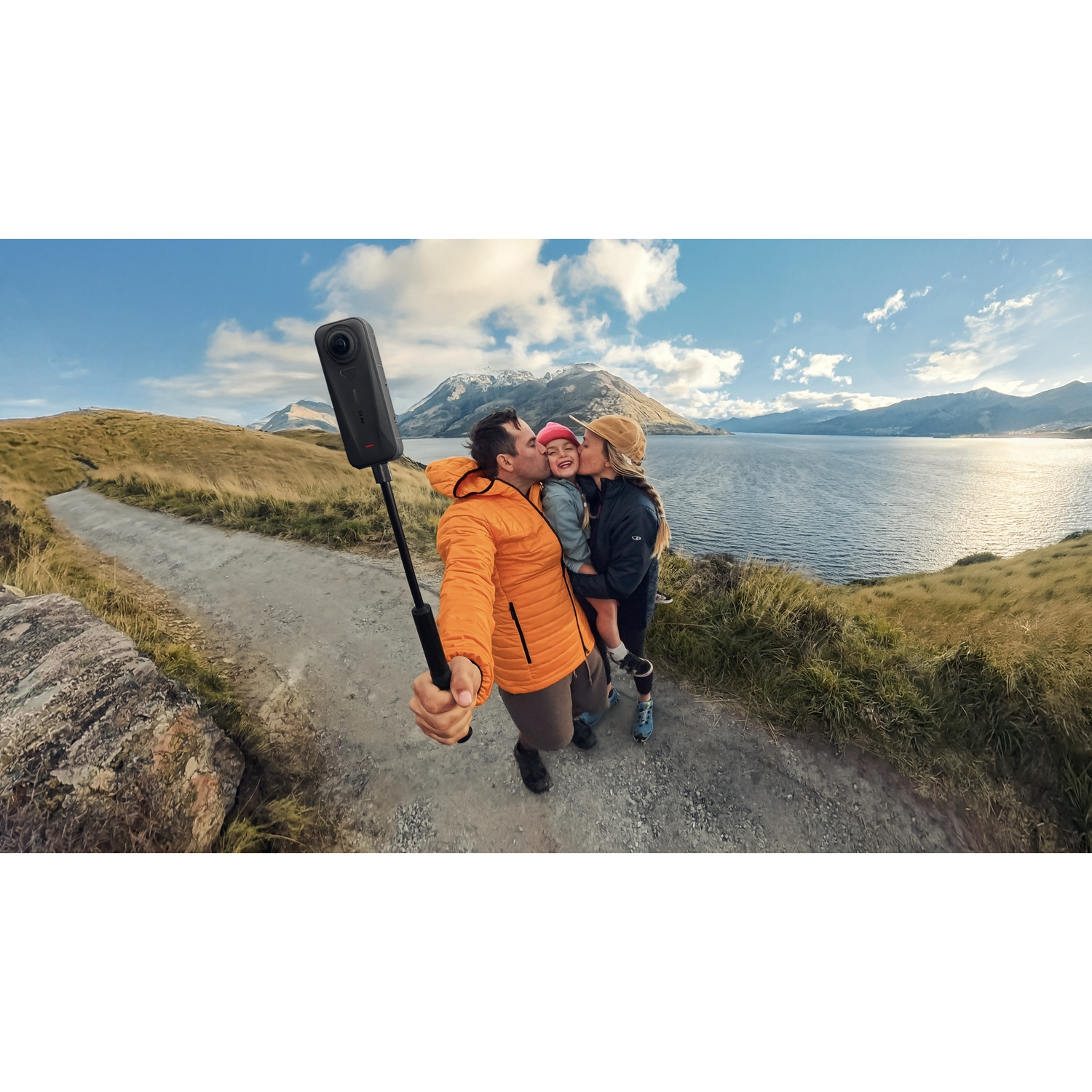 Selfie de famille avec une caméra 360. Un homme embrasse un enfant, une autre femme embrasse l'enfant. Montagnes et lac en arrière-plan.