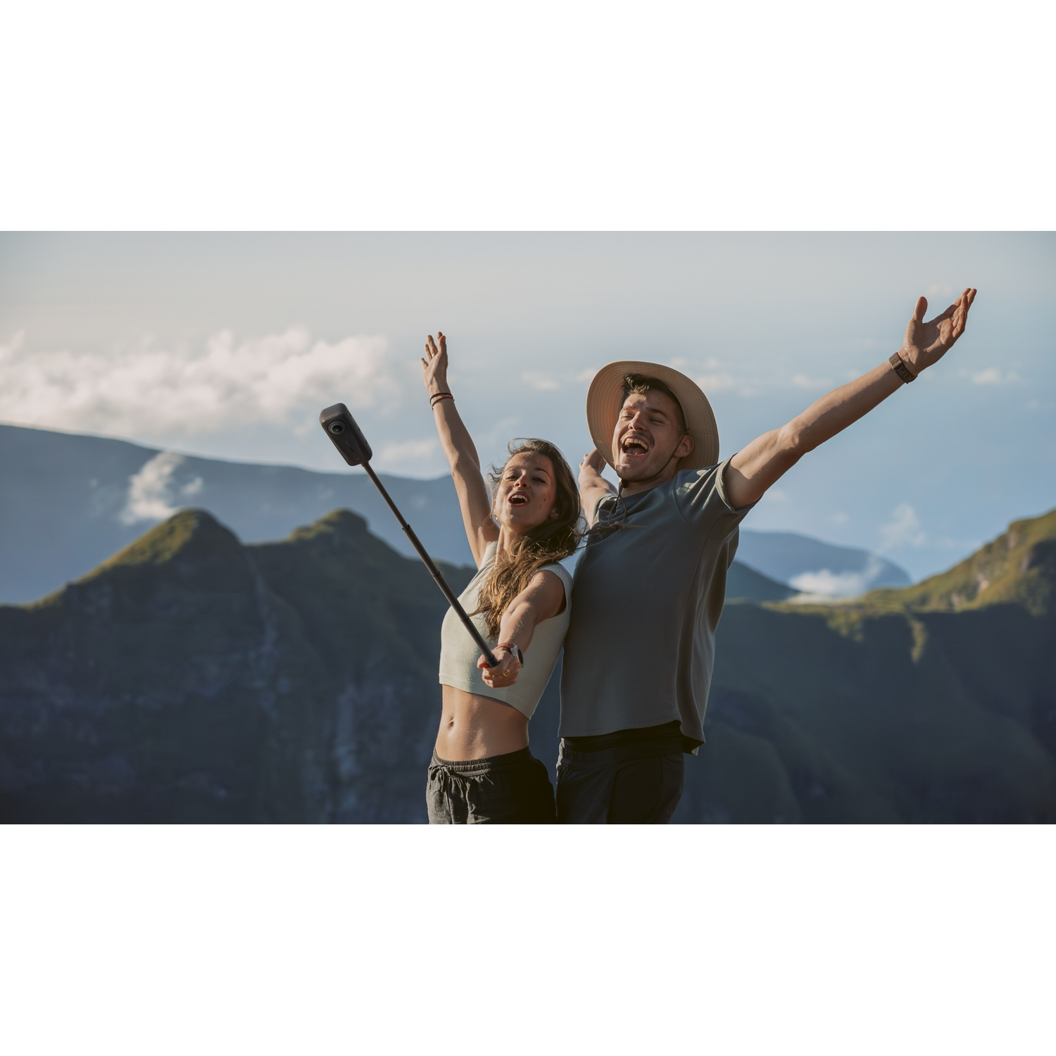 Un homme et une femme debout sur une montagne, les bras levés. L'homme porte un chapeau. Des montagnes en arrière-plan. Une perche à selfie est tenue.