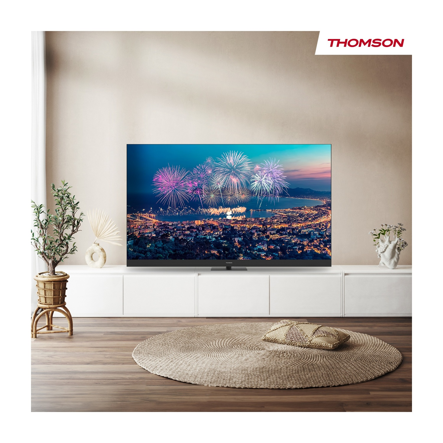 Soggiorno con TV che mostra fuochi d'artificio su una città. Tappeto e piante in primo piano. Logo Thomson in alto a destra.