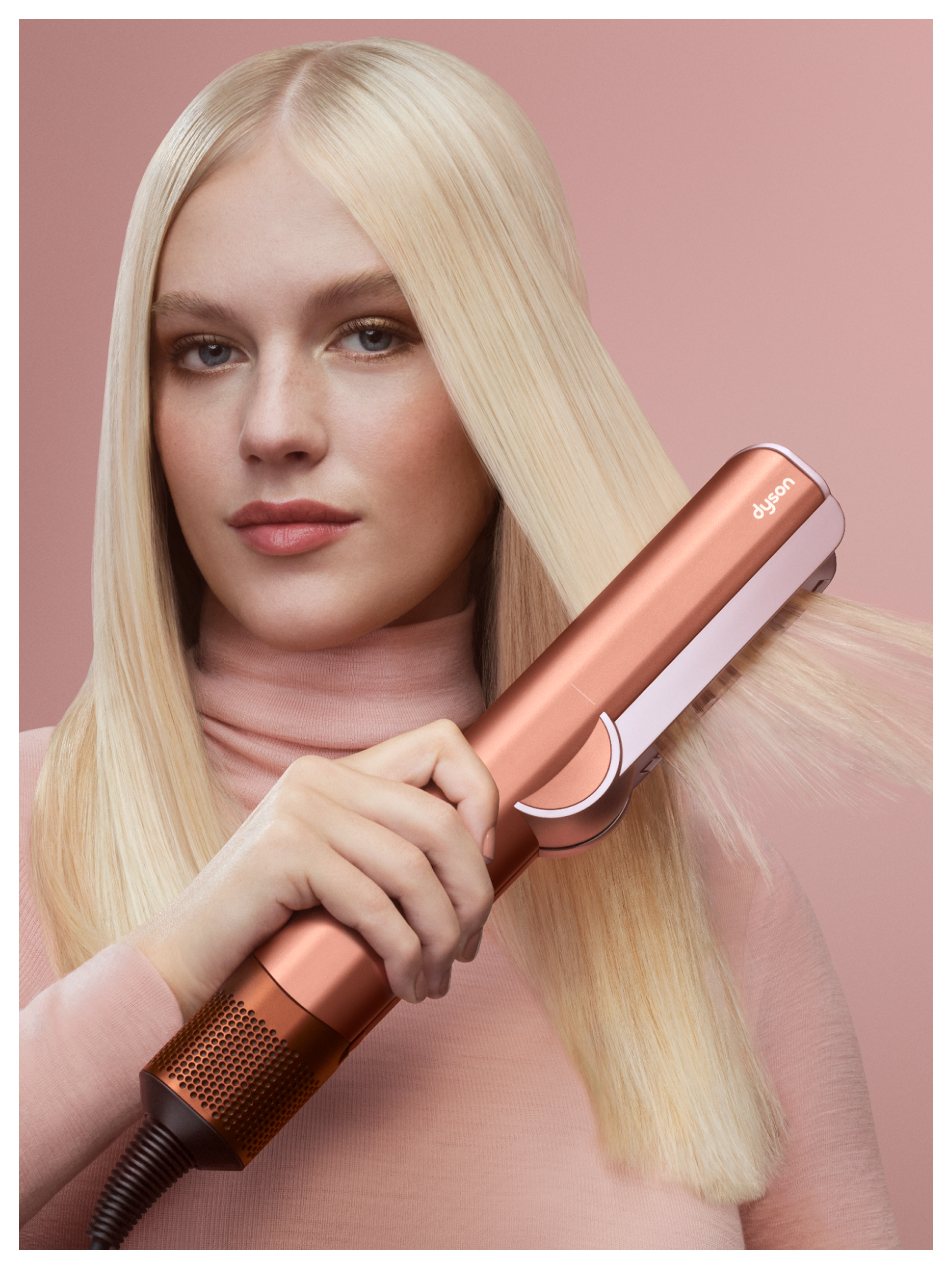 Mujer rubia usando una plancha de pelo Dyson. Lleva un top rosa. Fondo rosa. La plancha es de color cobre.