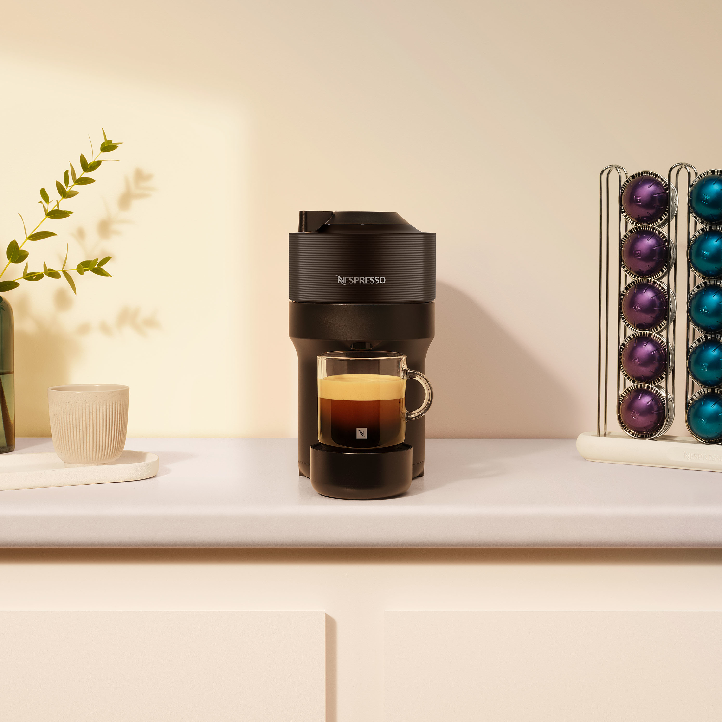 Una macchina Nespresso nera con una tazza di caffè, accanto a capsule e una pianta. Sfondo neutro.
