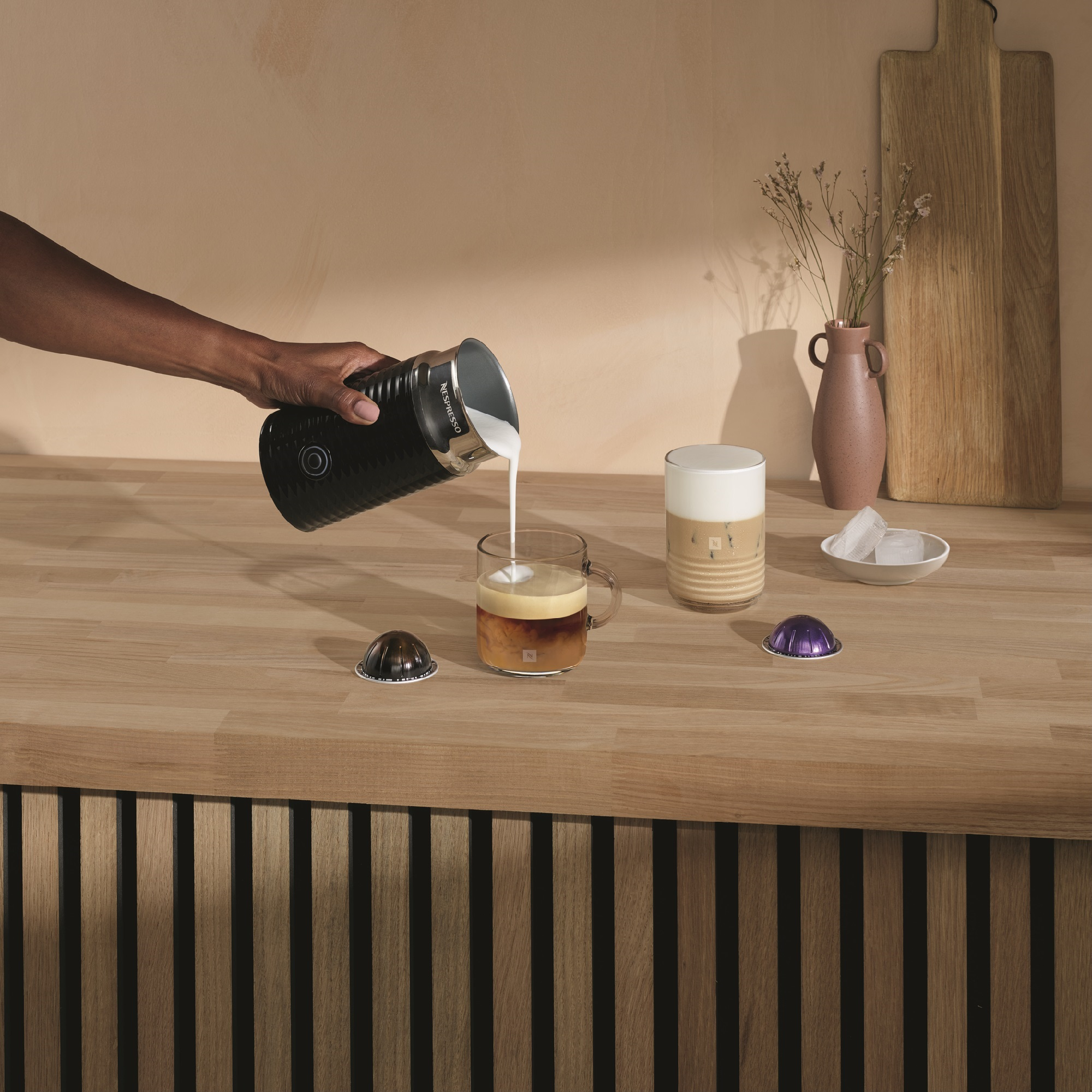 Una persona versa latte in una tazza di caffè. Altre tazze, capsule e ghiaccio su legno.