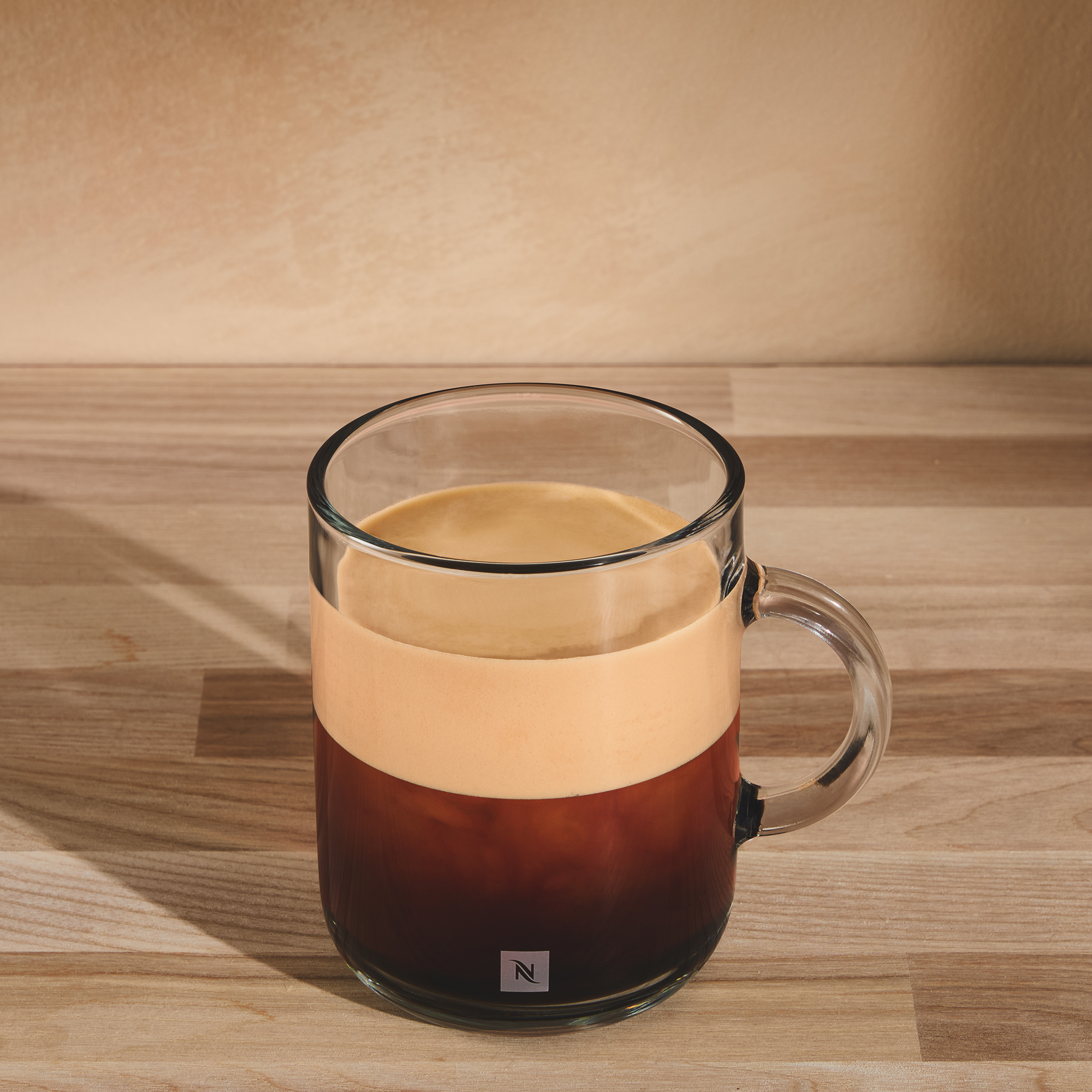Tazza di vetro trasparente con strati di caffè su superficie in legno. Parete marrone chiaro sullo sfondo.