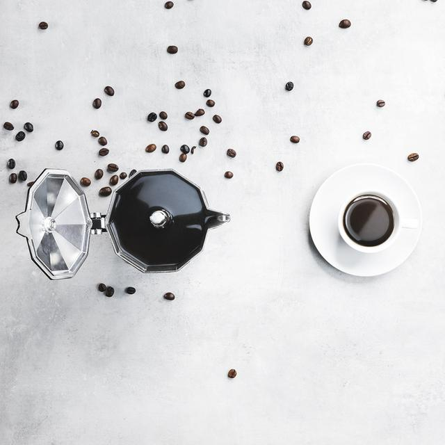 Vista aérea de granos de café esparcidos alrededor de una cafetera y una taza de café sobre una superficie blanca.