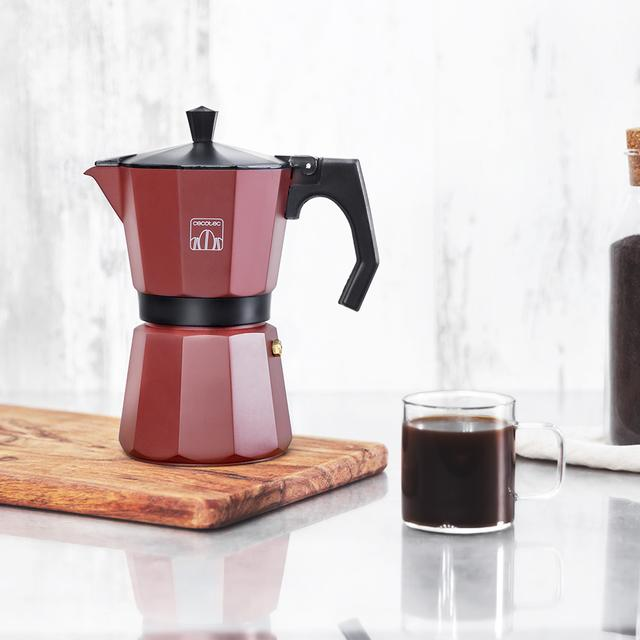 Una cafetera roja está sobre una tabla de madera. Al lado, una taza de vidrio contiene café oscuro. En el fondo, una botella y una superficie blanca.